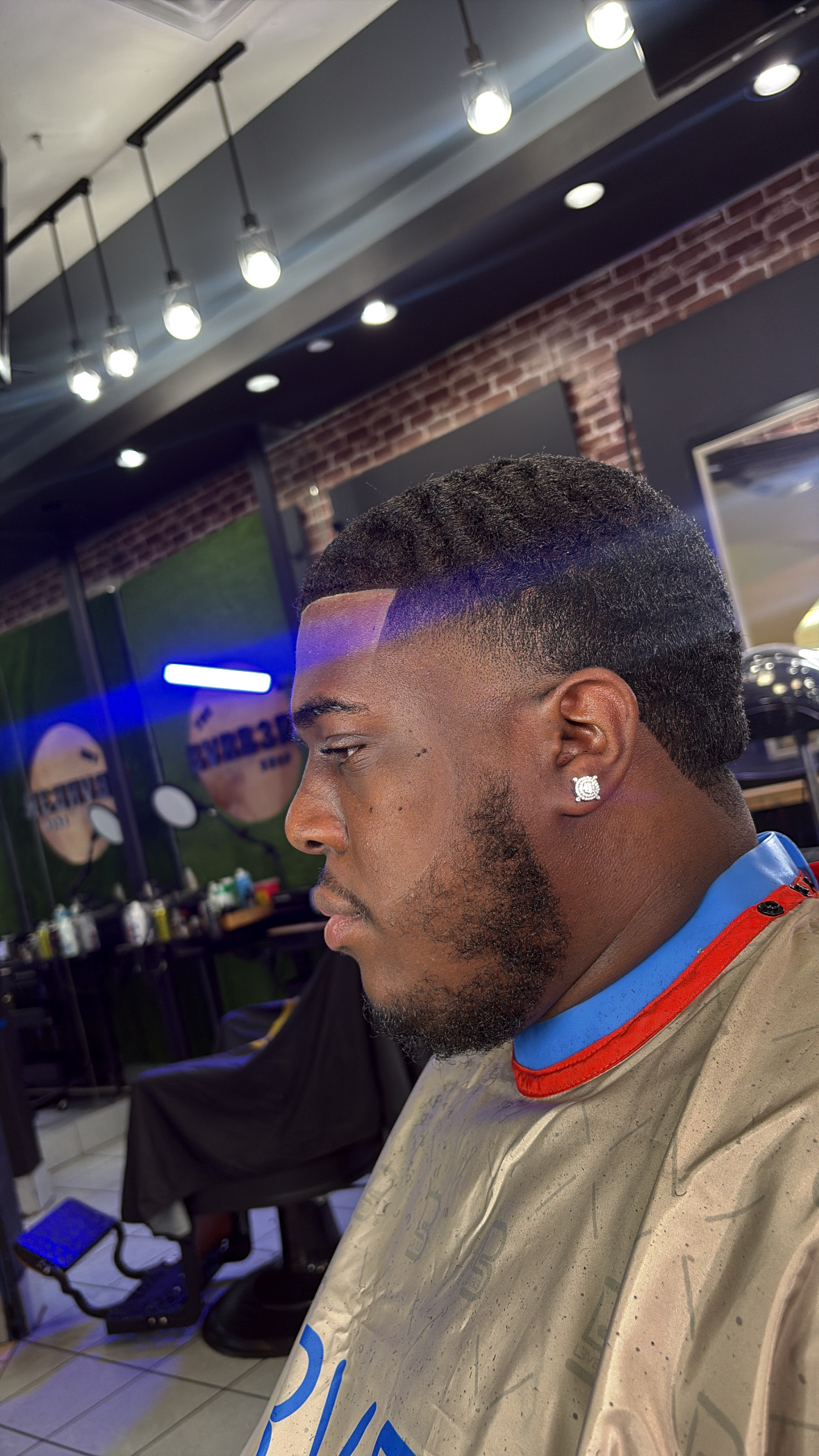 Side profile of a young man with a fade haircut, beard, and diamond earring in a barbershop or salon.