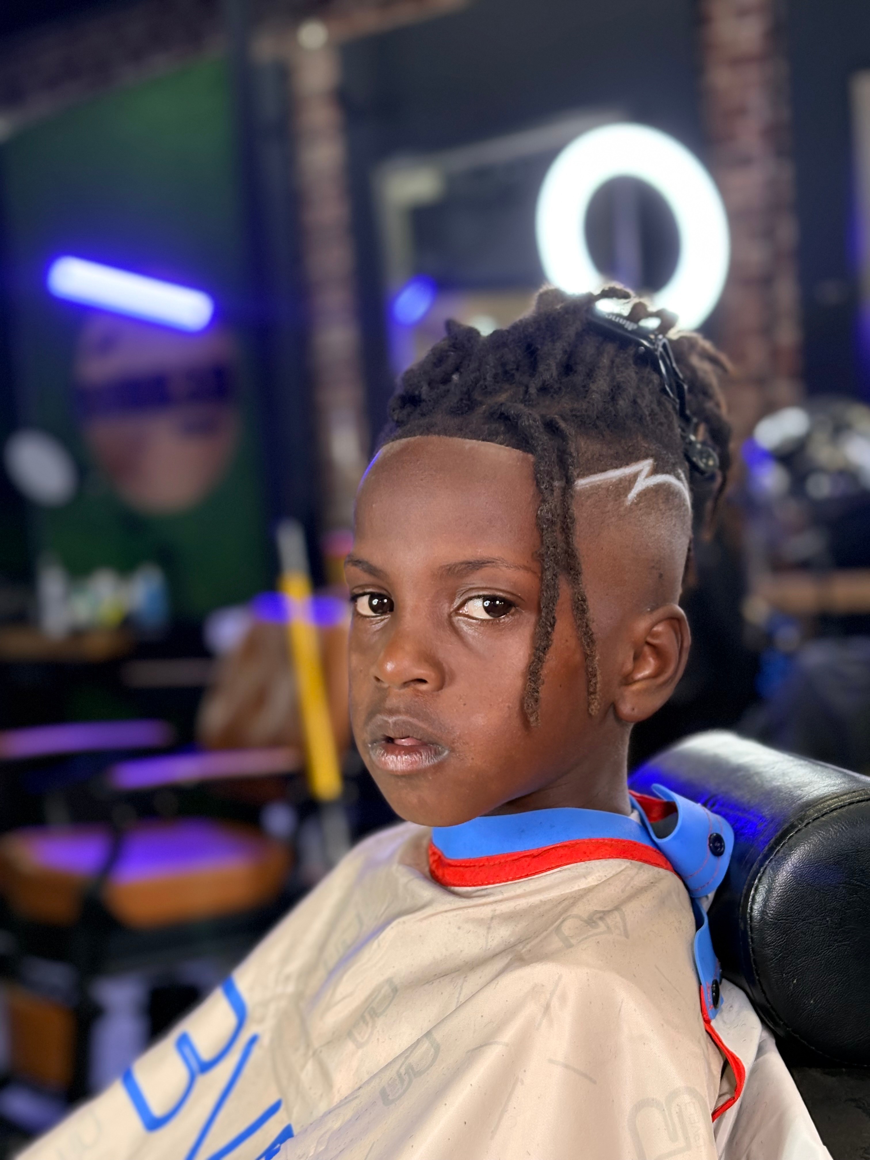 Child with styled hair and a barber cape in a barbershop, illuminated by neon lights, looking at the camera.