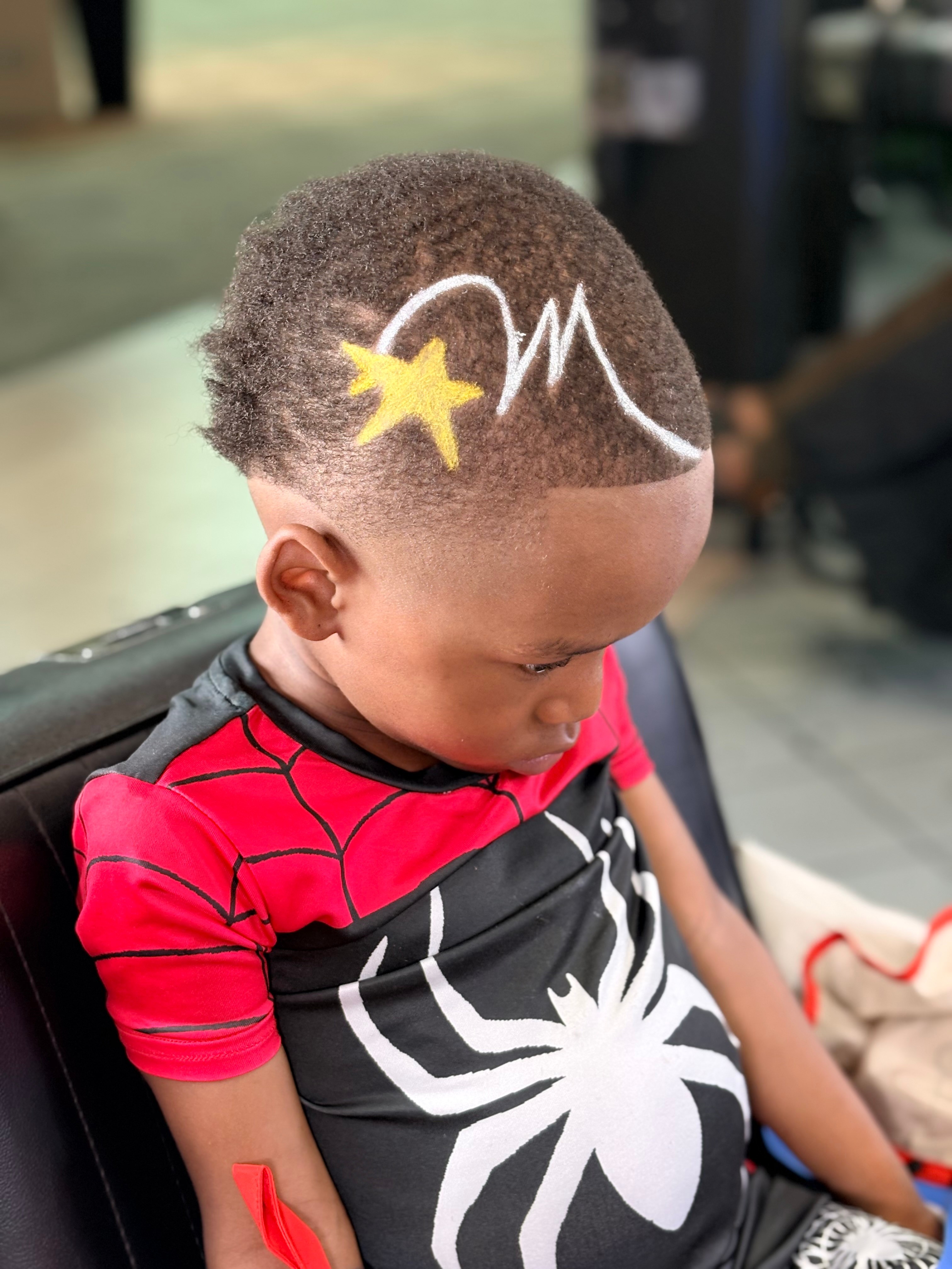Young child with a star and wave design shaved into hair, wearing a Spider-Man shirt, looking down.