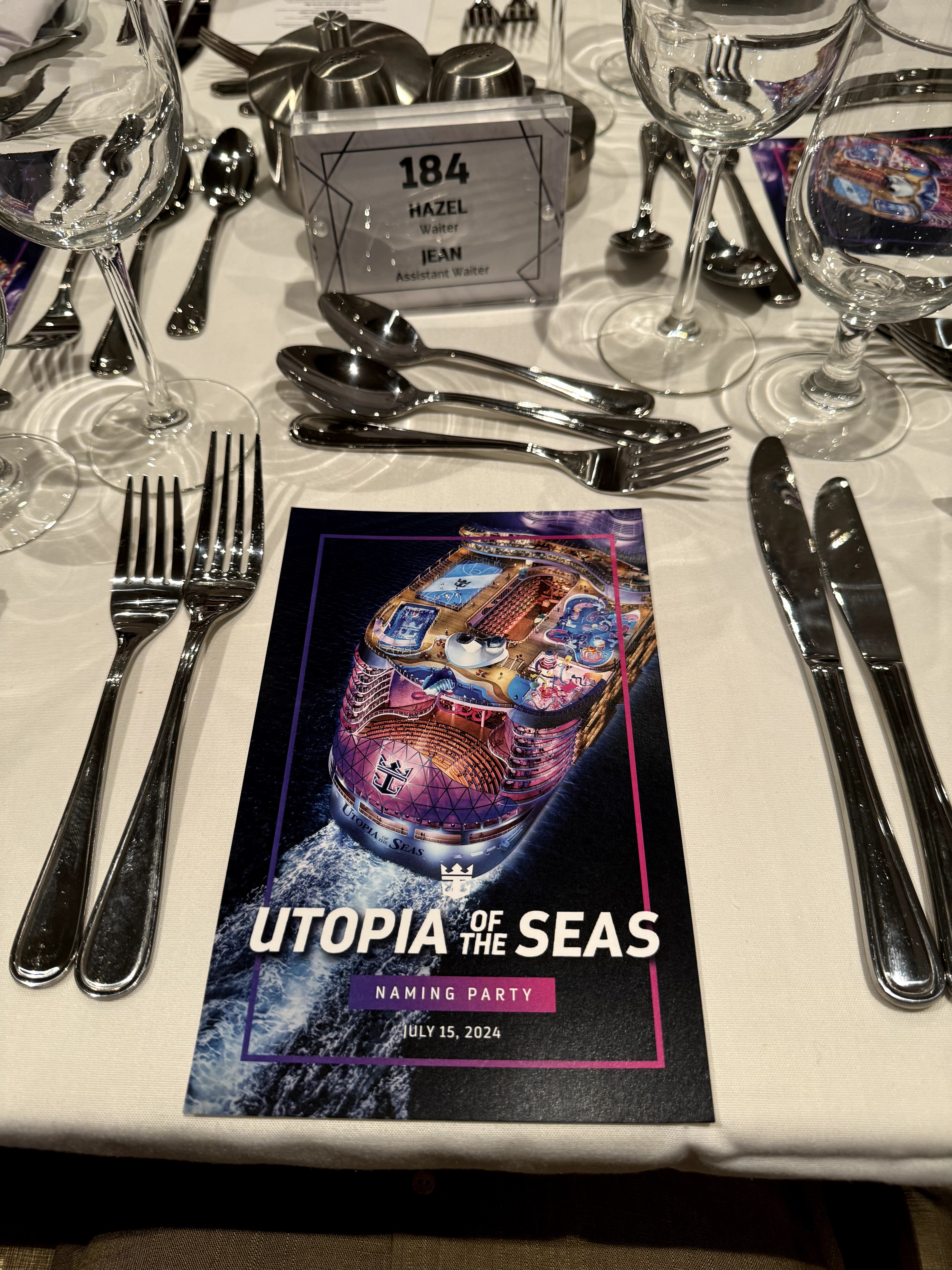 Table setting with glasses, cutlery, and a program for the Utopia of the Seas naming party, dated May 15, 2024.