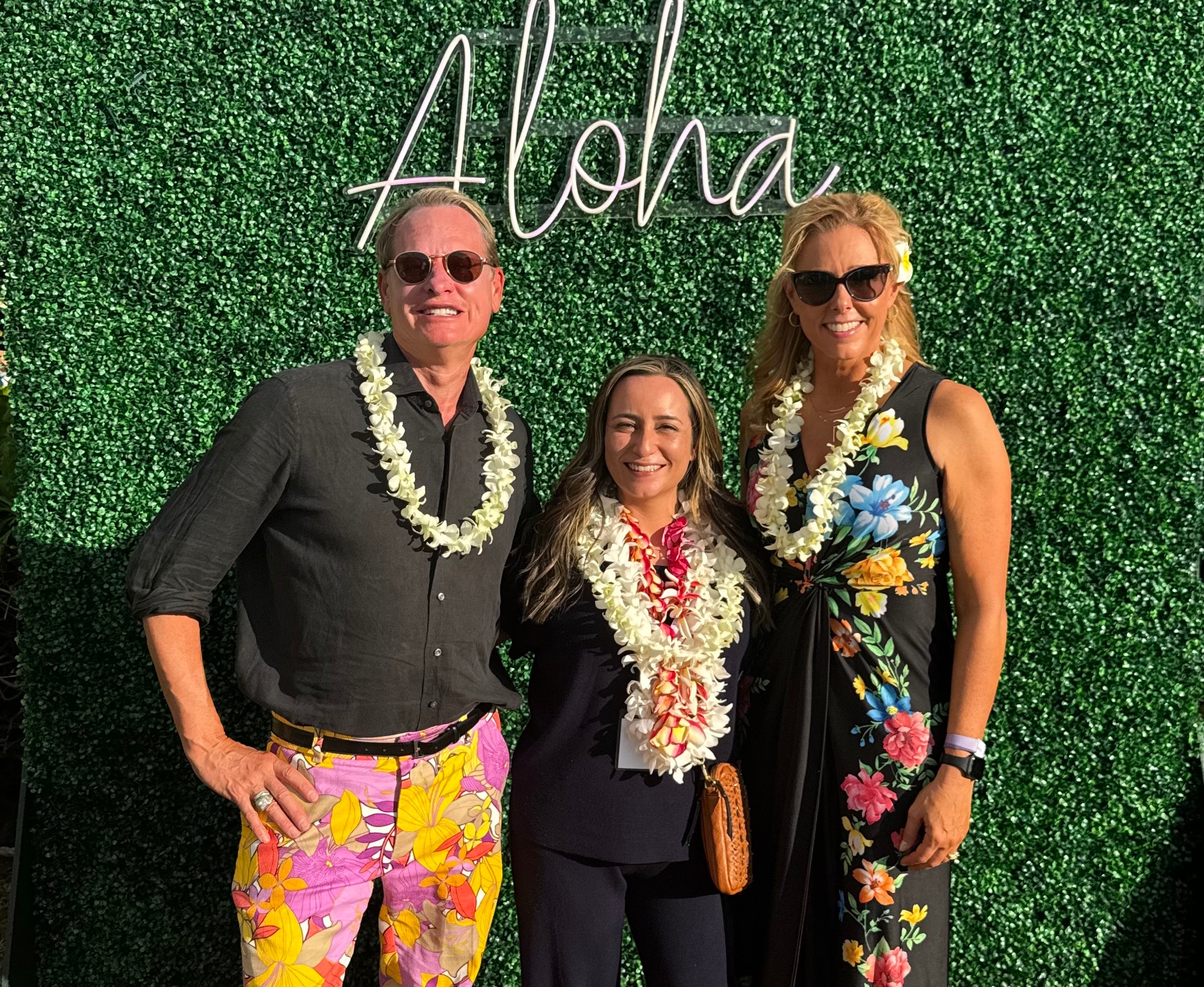 Three people smiling in front of a green hedge with a neon 'Aloha' sign, wearing leis and tropical clothing.