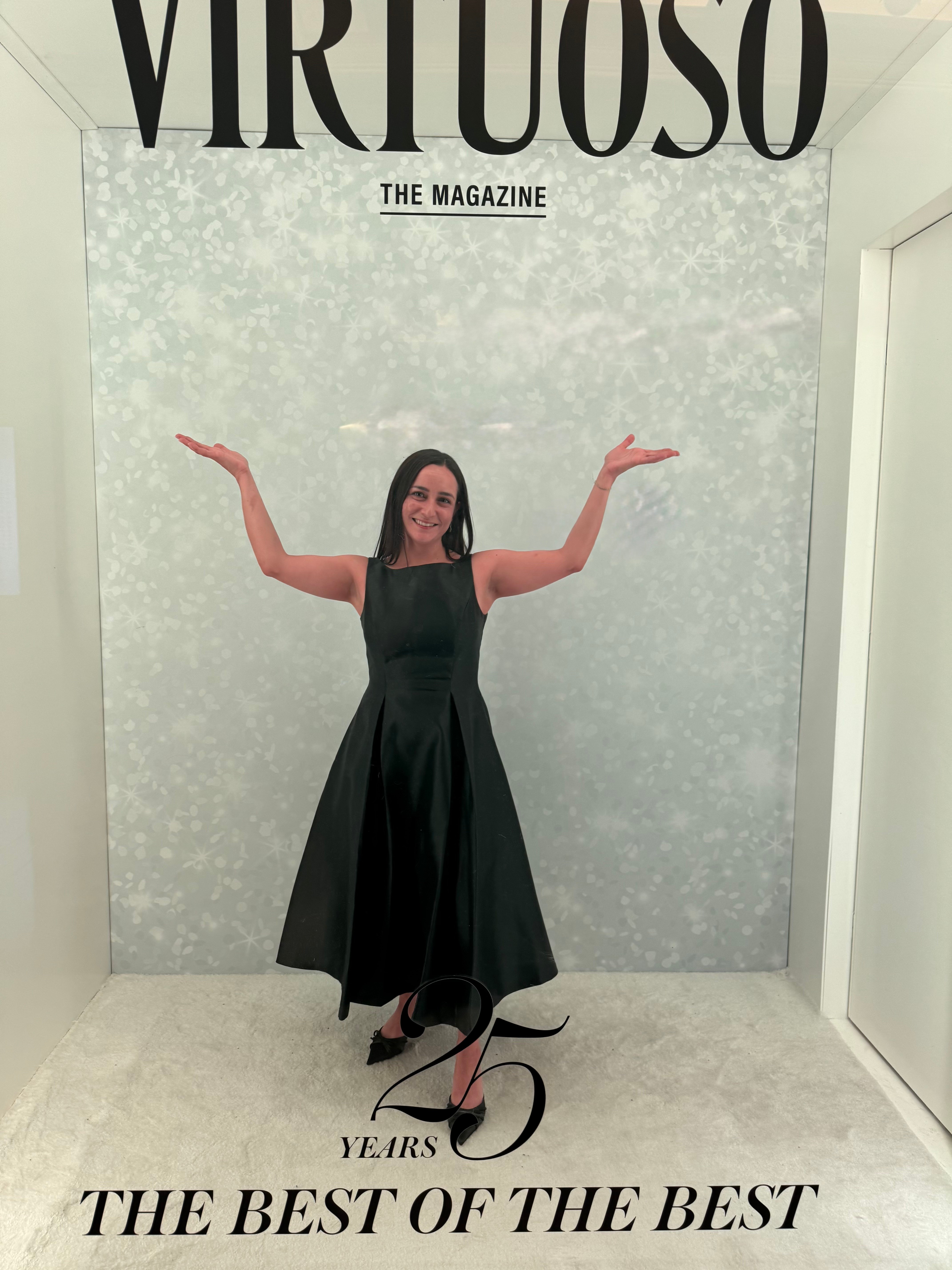 Woman in a black dress standing with arms outstretched in front of a backdrop with text, celebrating 25 years.