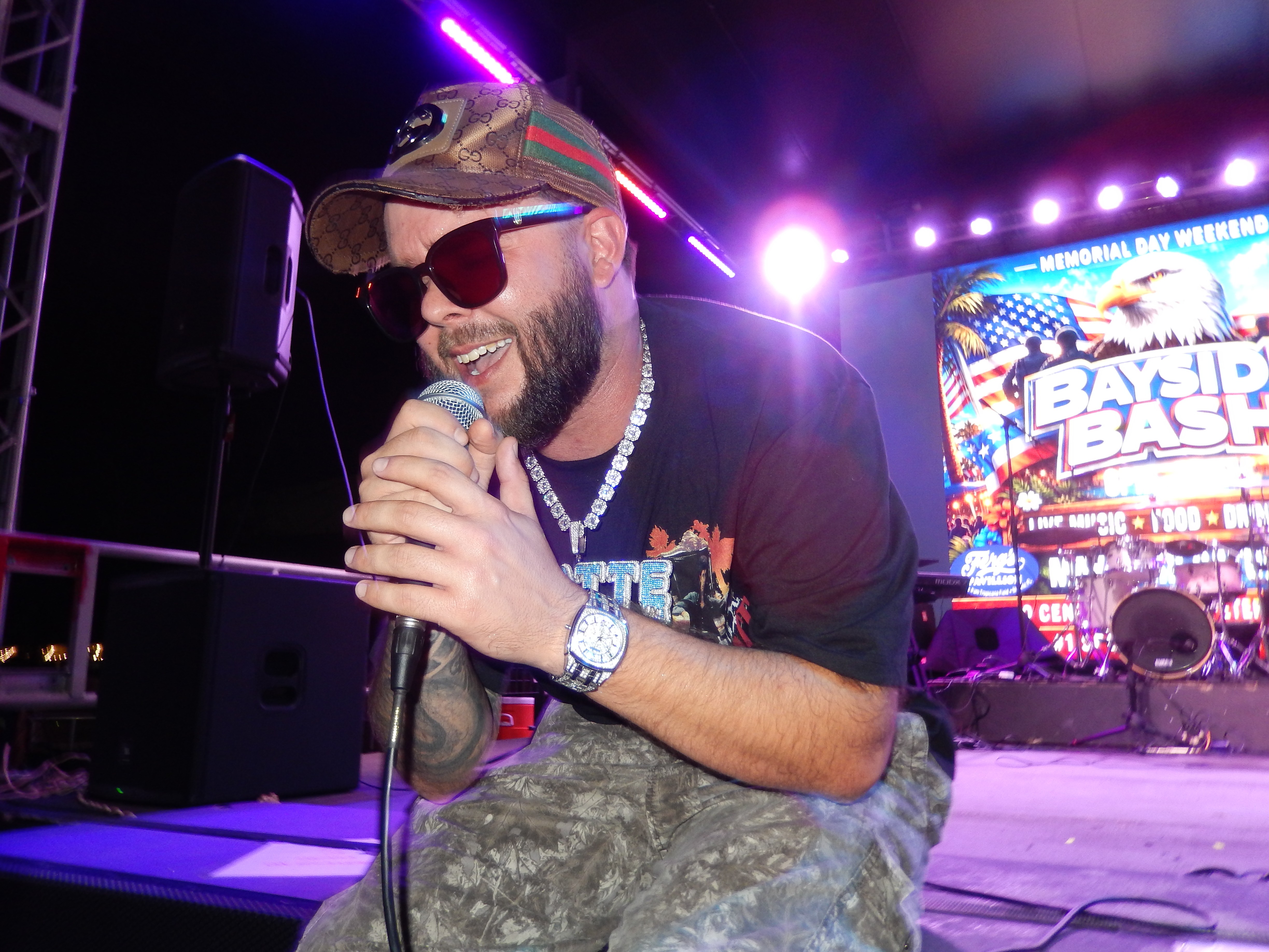 Man with sunglasses, cap, and camouflage pants holding a microphone on stage with colorful lights and a screen in background.