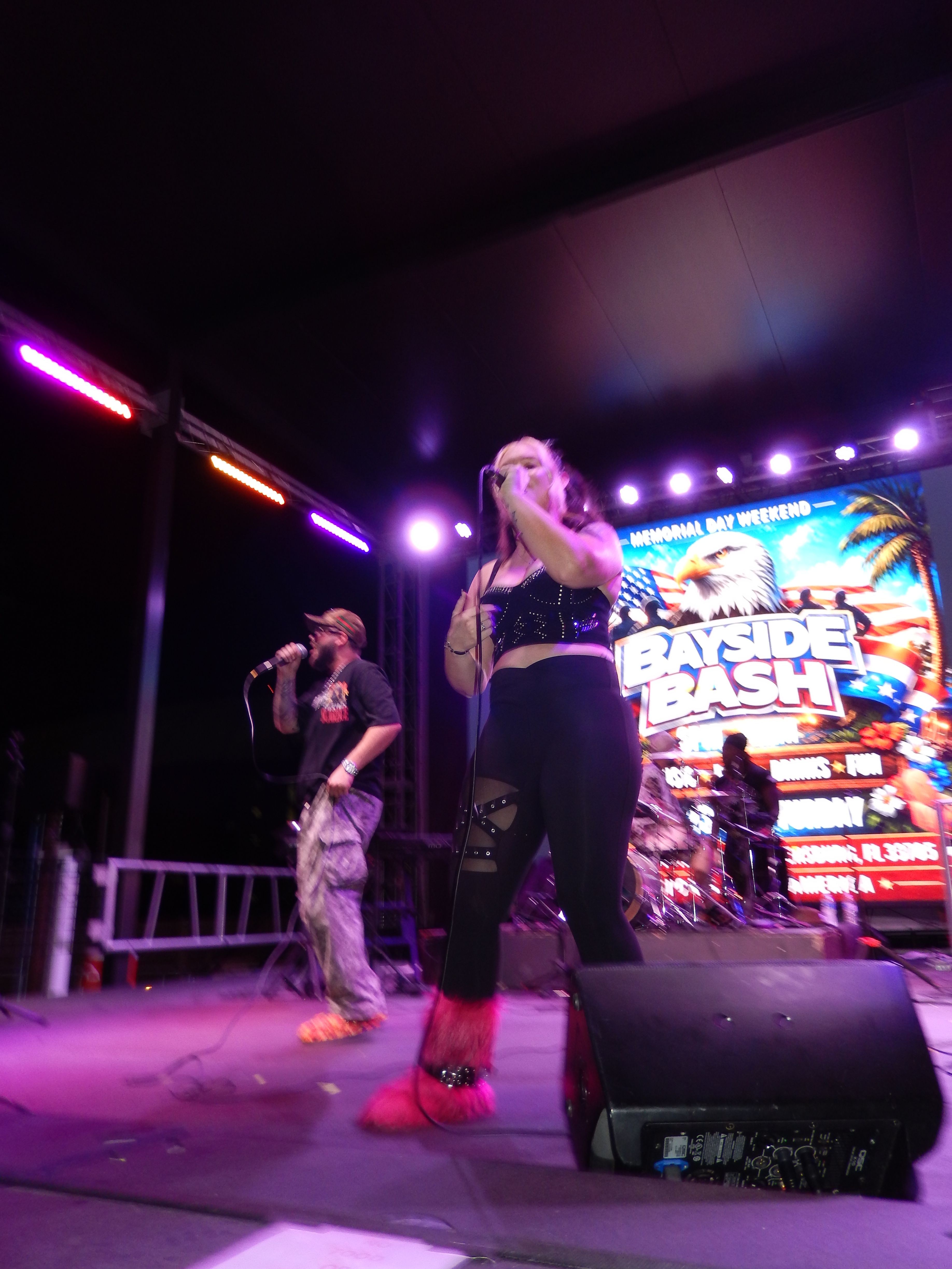 Two performers on stage with colorful background, one singing into a microphone, the other standing nearby, stage lighting visible.