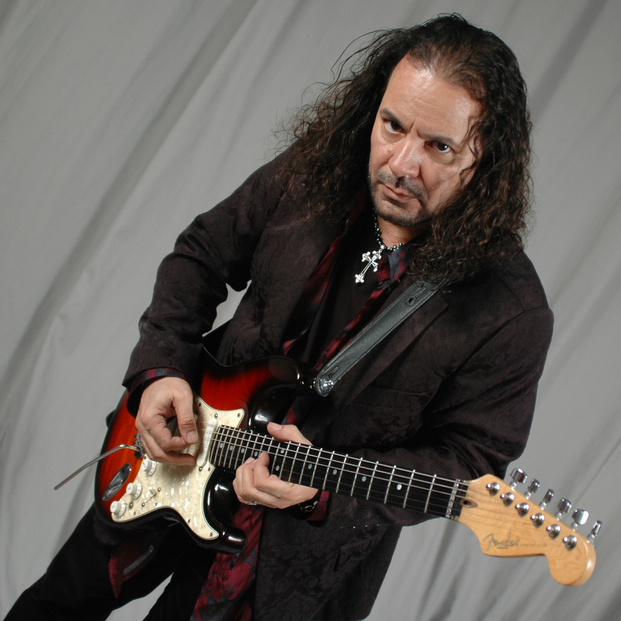Man with long curly hair playing an electric guitar, wearing a dark jacket, against a plain background.