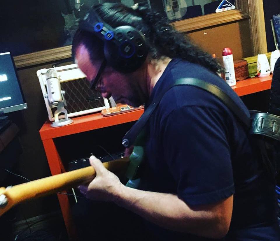 Person with long curly hair wearing headphones playing guitar in a recording studio.