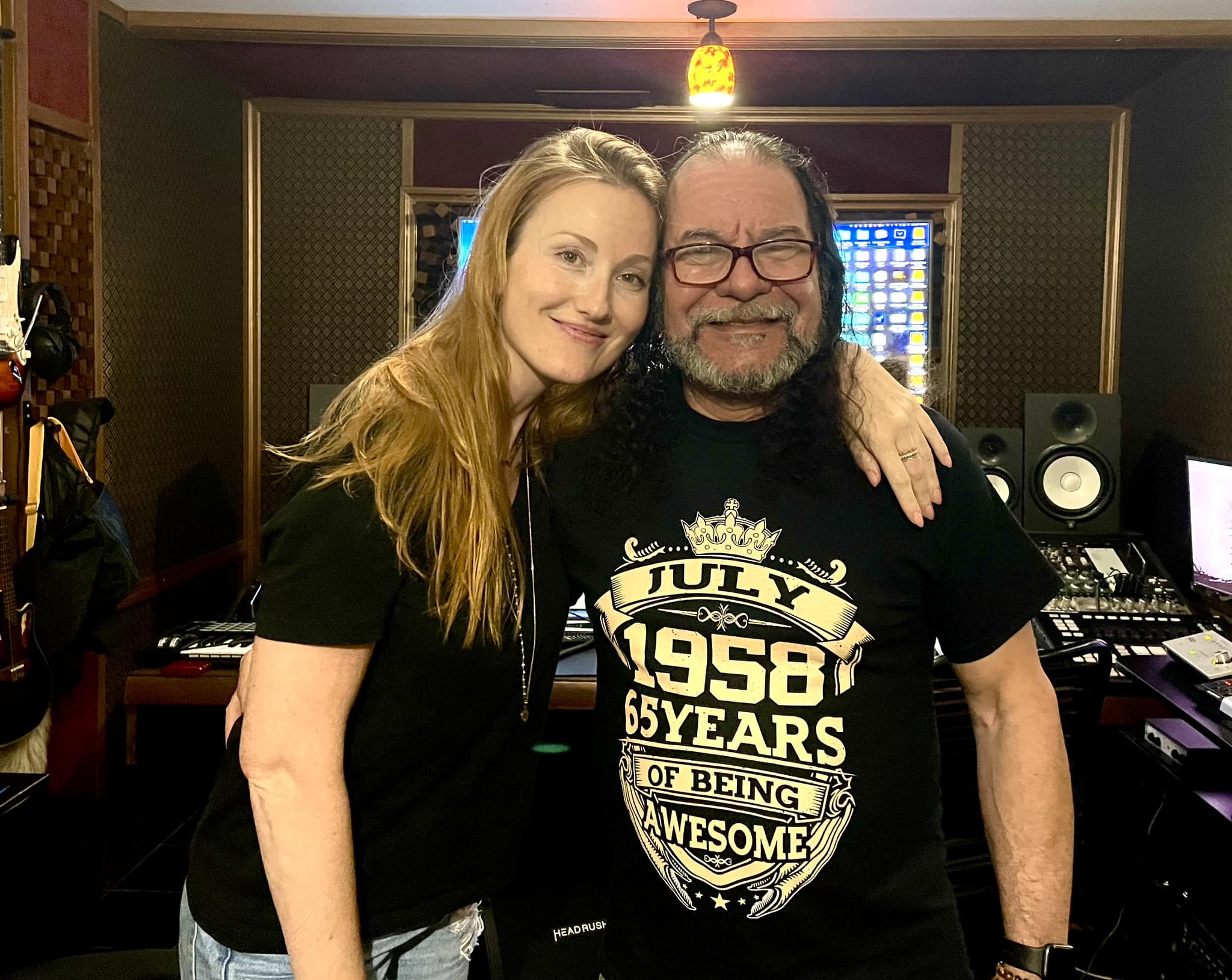 Two people smiling, one with long blonde hair and the other with long dark hair and glasses, in a recording studio.