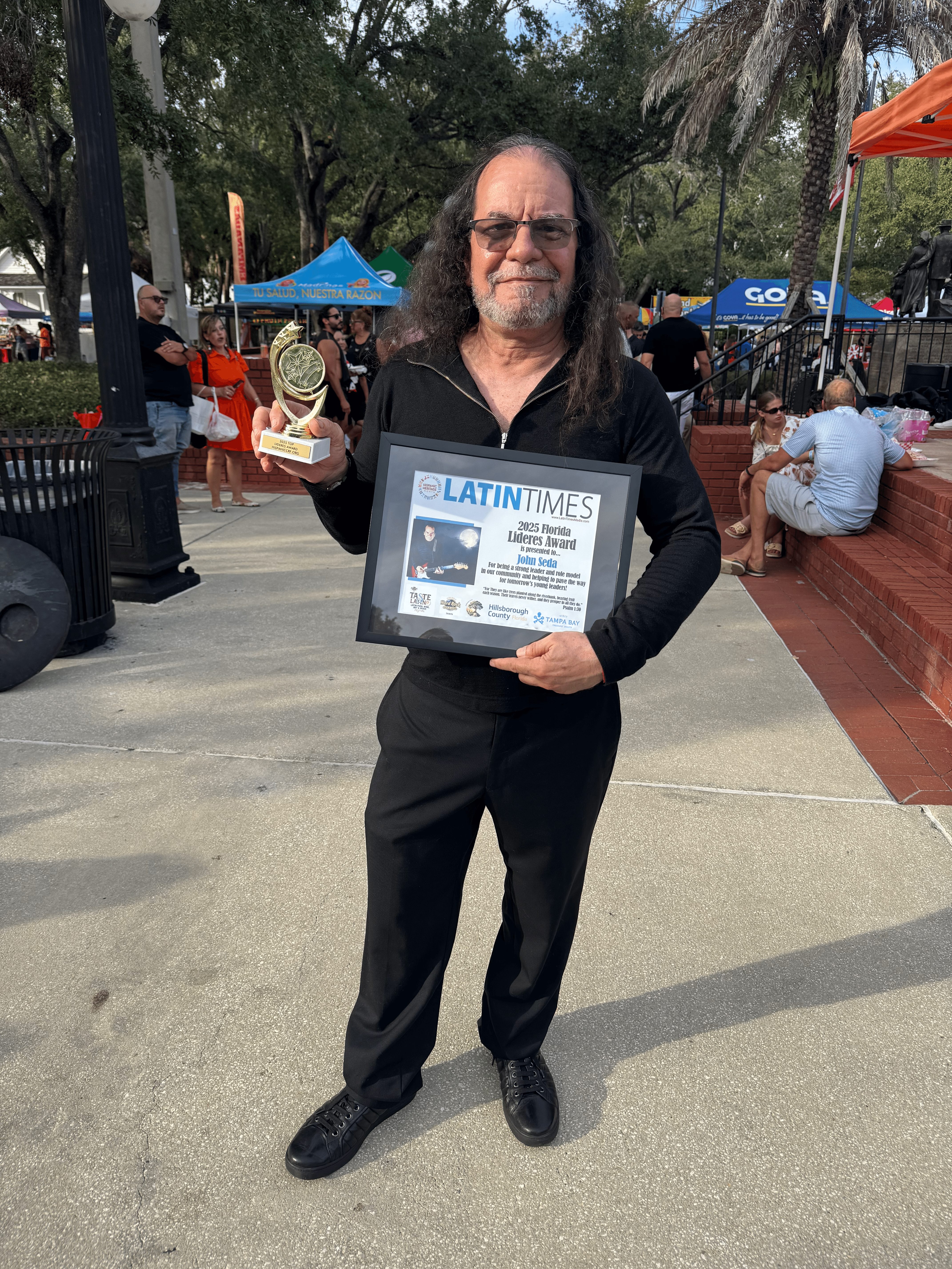 Man with long hair and glasses holding a framed certificate outdoors at a park event.