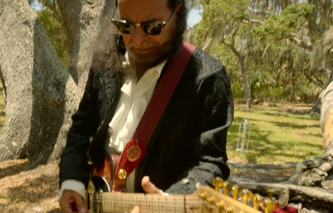 Person wearing sunglasses playing guitar outdoors with trees and grass in background.