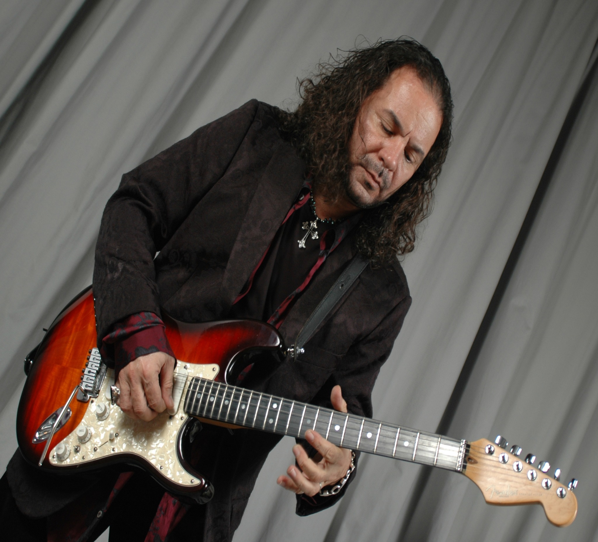 Person playing an electric guitar with long curly hair, wearing a dark jacket, in front of a curtain backdrop.