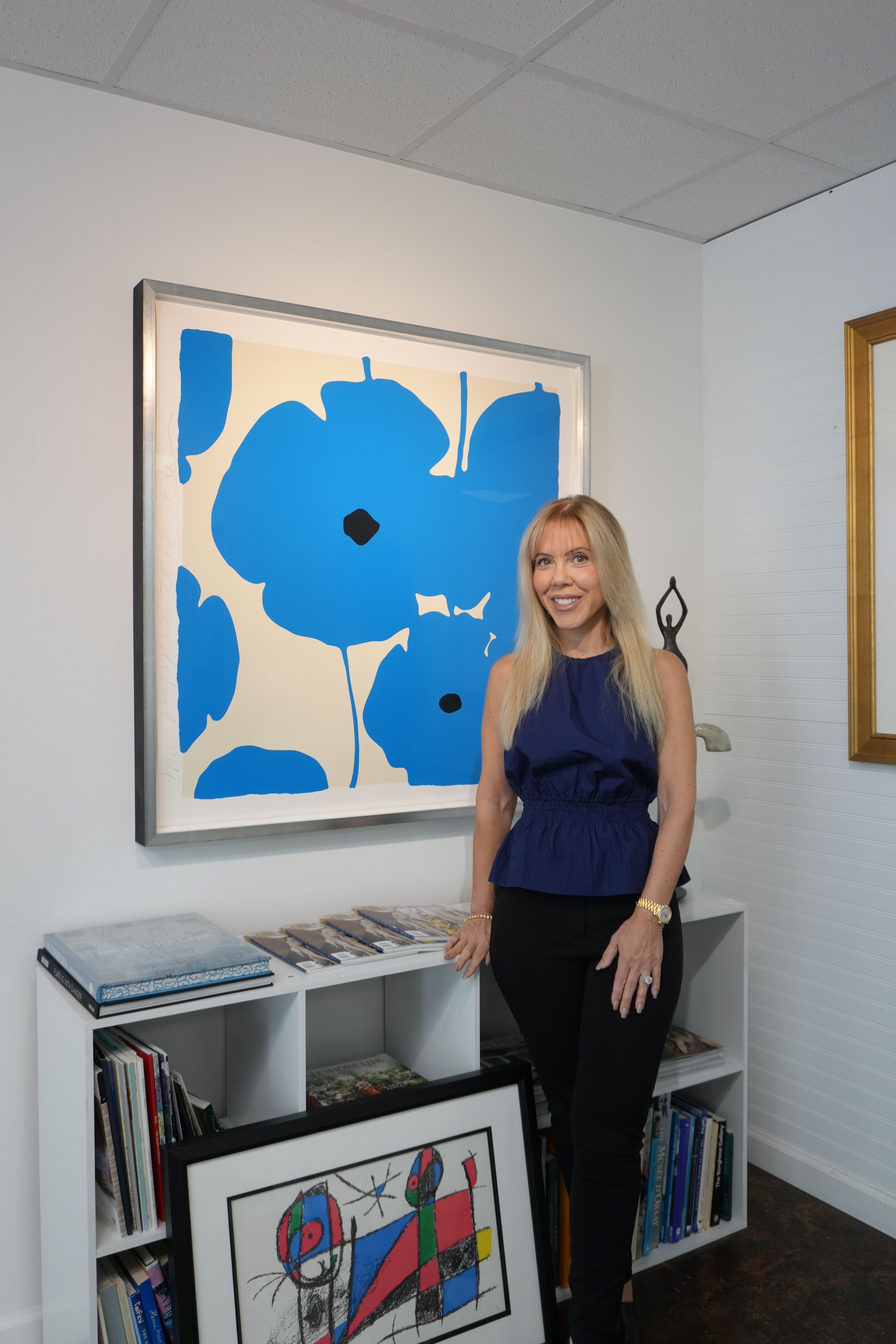 Woman with long blonde hair wearing a blue top and black pants standing next to a white bookshelf with artwork and books, in a room with white walls and tiled floor.