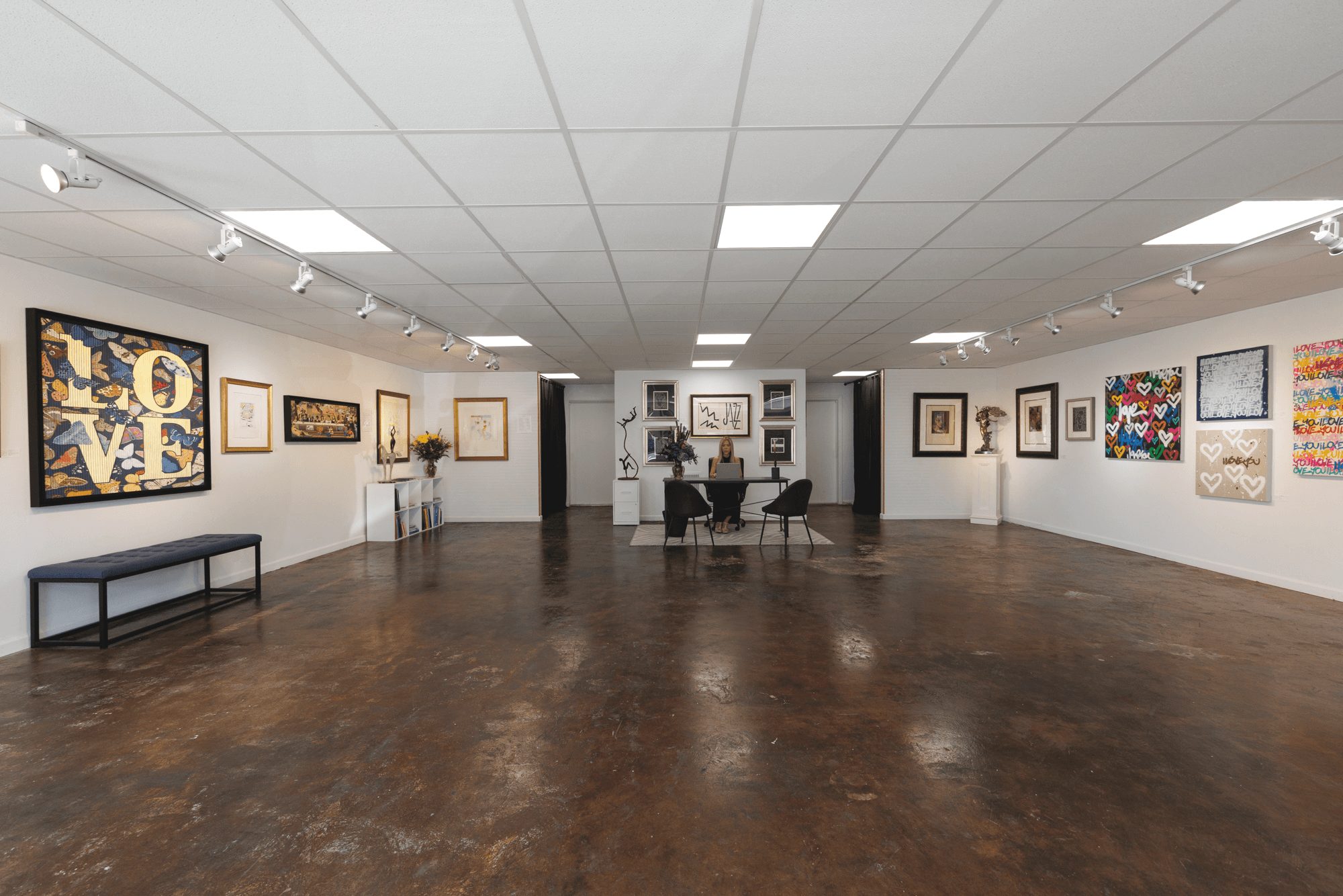 Art gallery with white walls, framed paintings, and a table with chairs in the center, wooden floor, ceiling lights, and a bench.