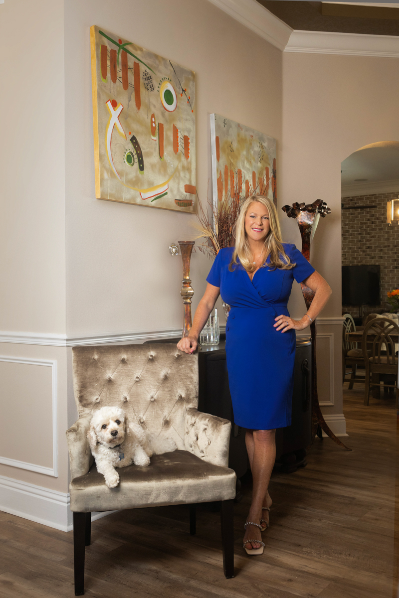 Woman in blue dress standing next to a tufted bench with a small dog on it, in a decorated living room.