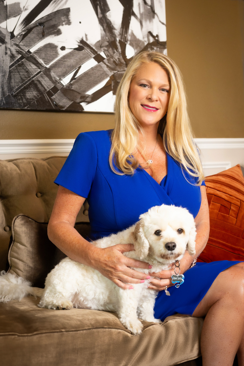 Woman in blue dress sitting on sofa with white dog, abstract artwork on wall behind, warm indoor setting.