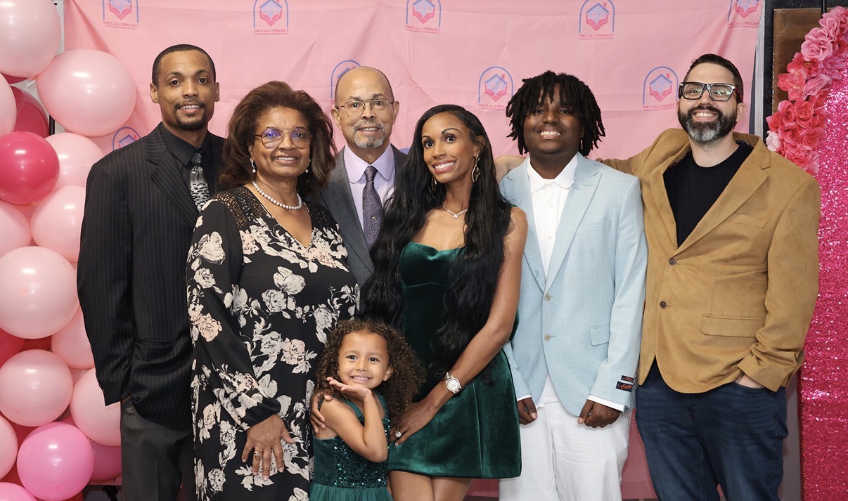 Group of seven people standing together indoors, pink background with balloons and flowers, smiling for the photo.