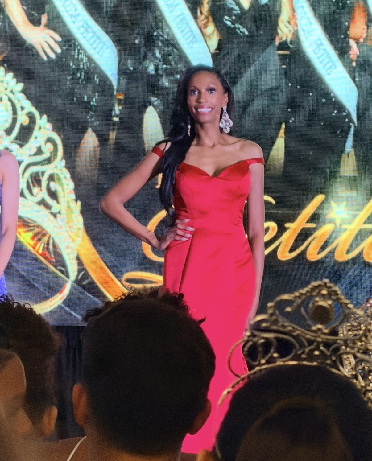 Woman in a red gown on stage with a crown in the foreground, background features a large screen with people and text.