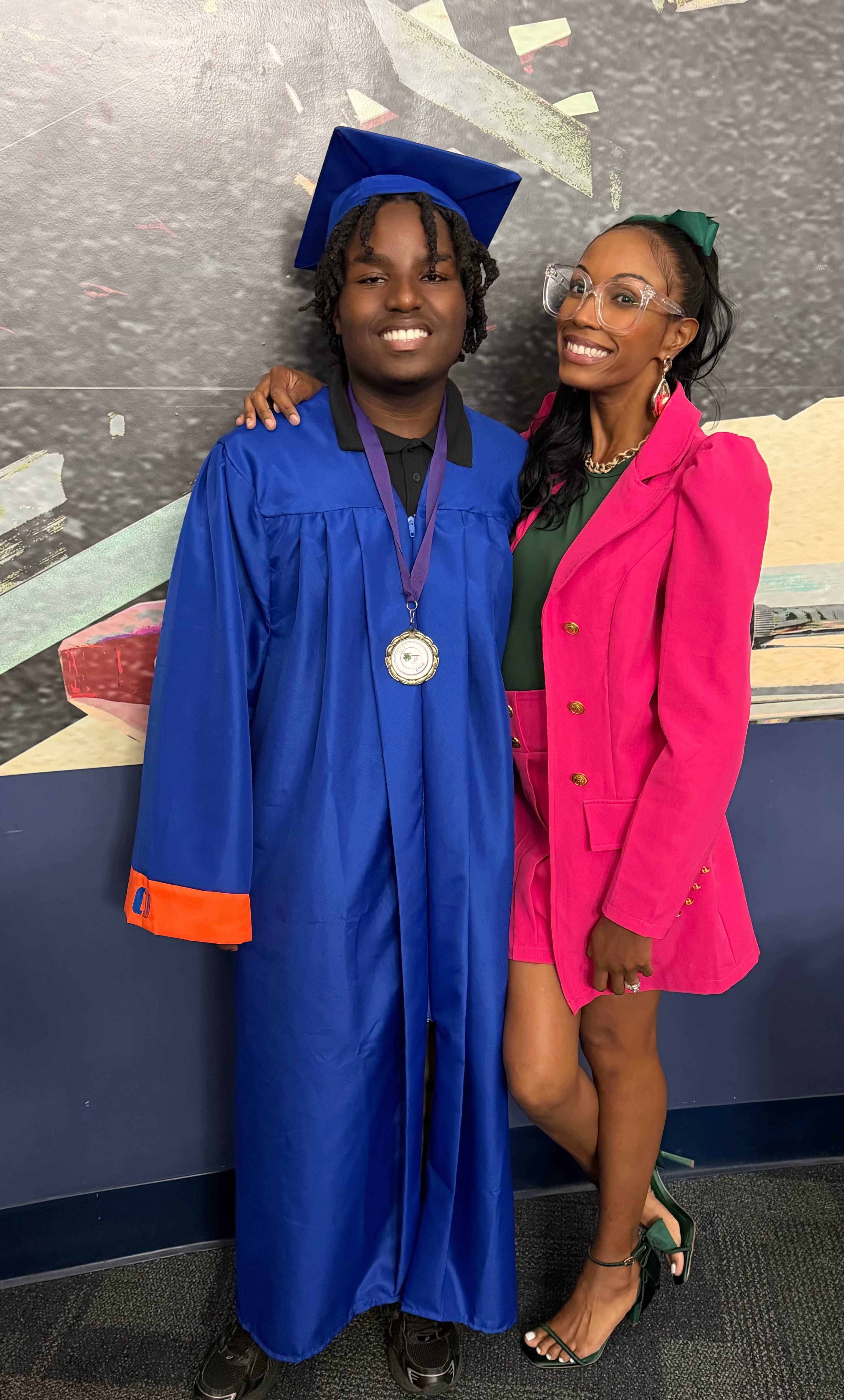 Two women smiling, one in a blue graduation gown and cap, the other in a pink blazer and glasses, standing together.