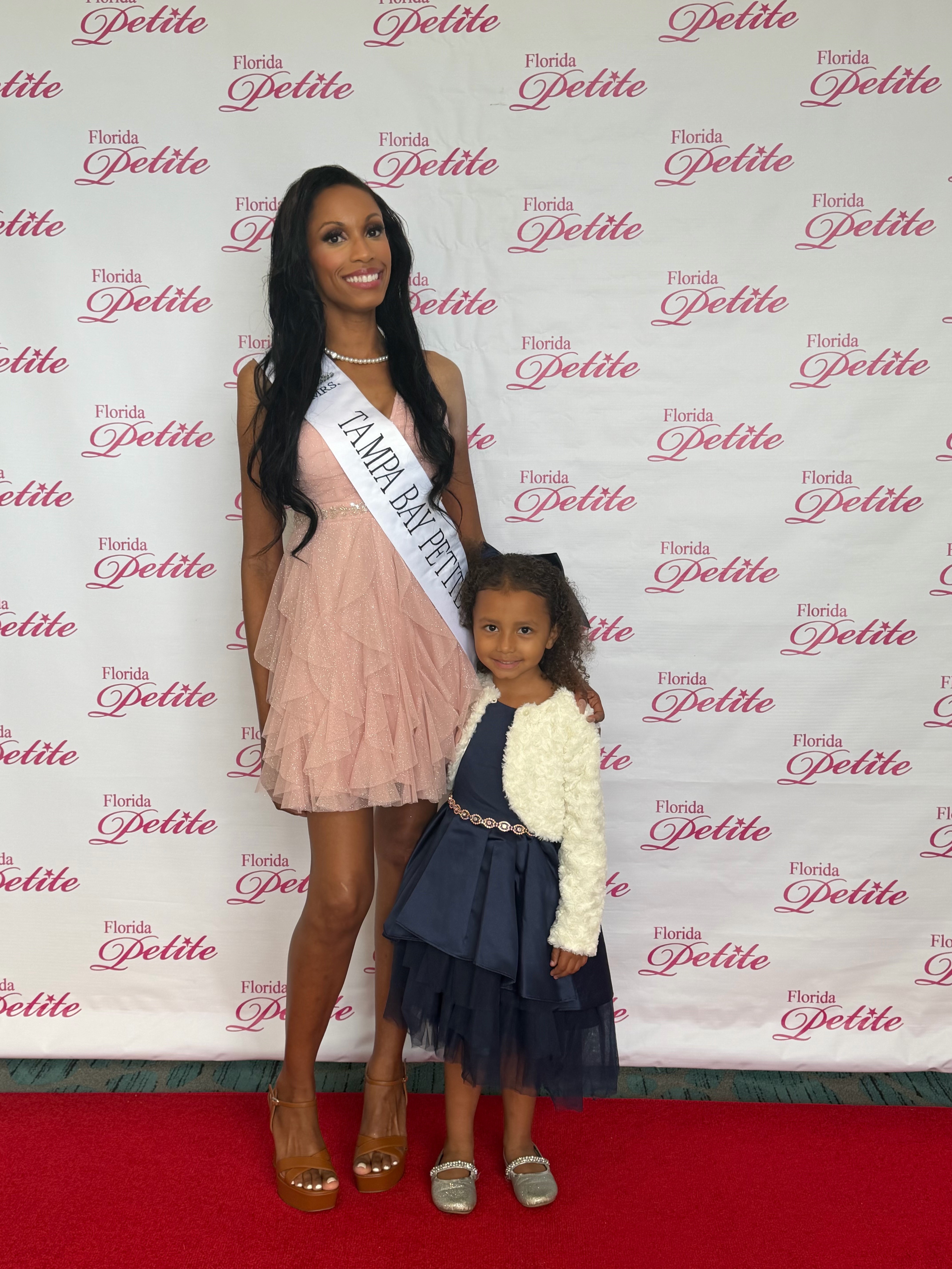 Woman and young girl pose in front of a backdrop with pink text, woman wears a sash, both smile, red carpet below.