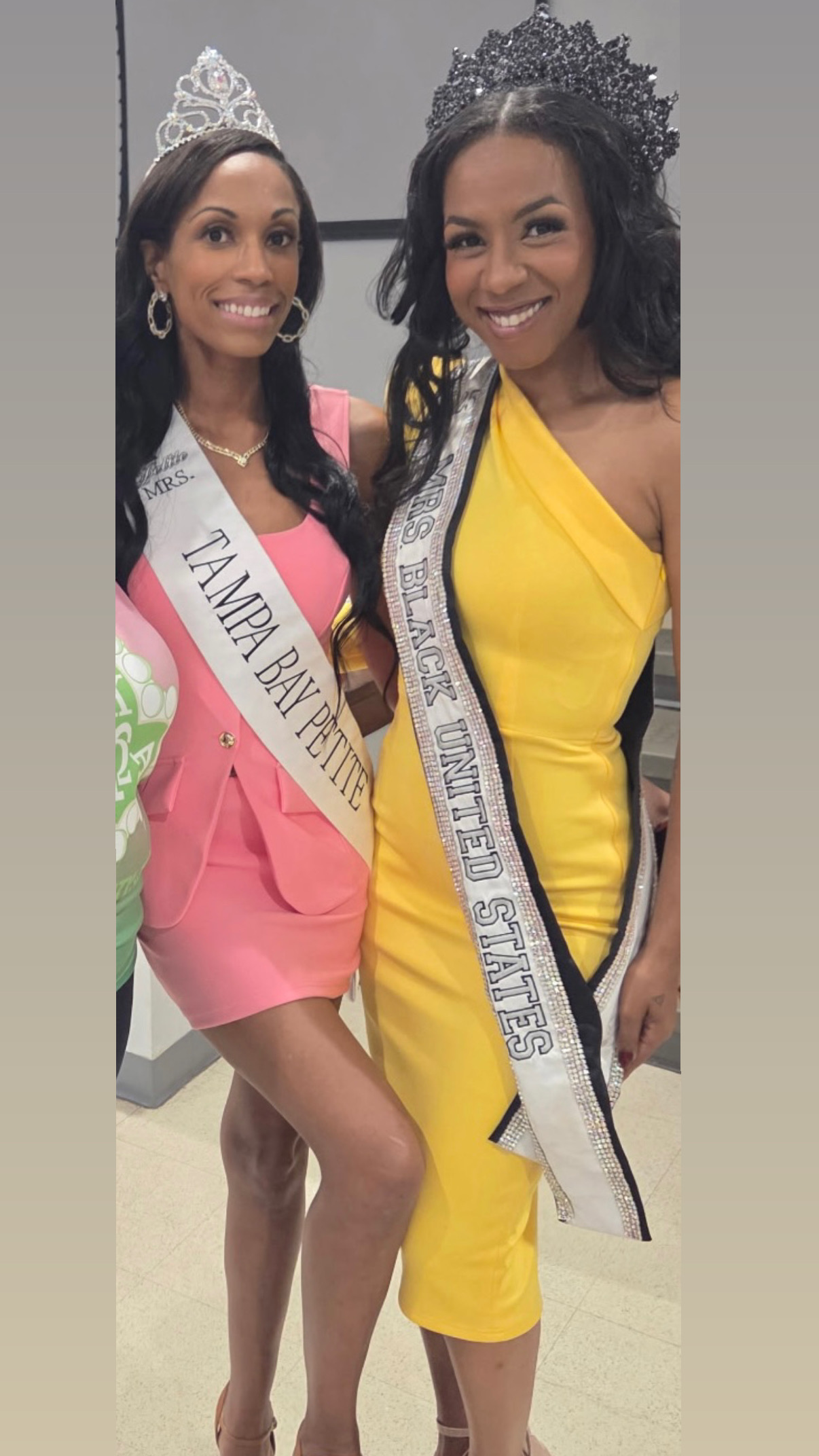Two women wearing crowns and sashes, smiling, one in pink and the other in yellow, standing together indoors.