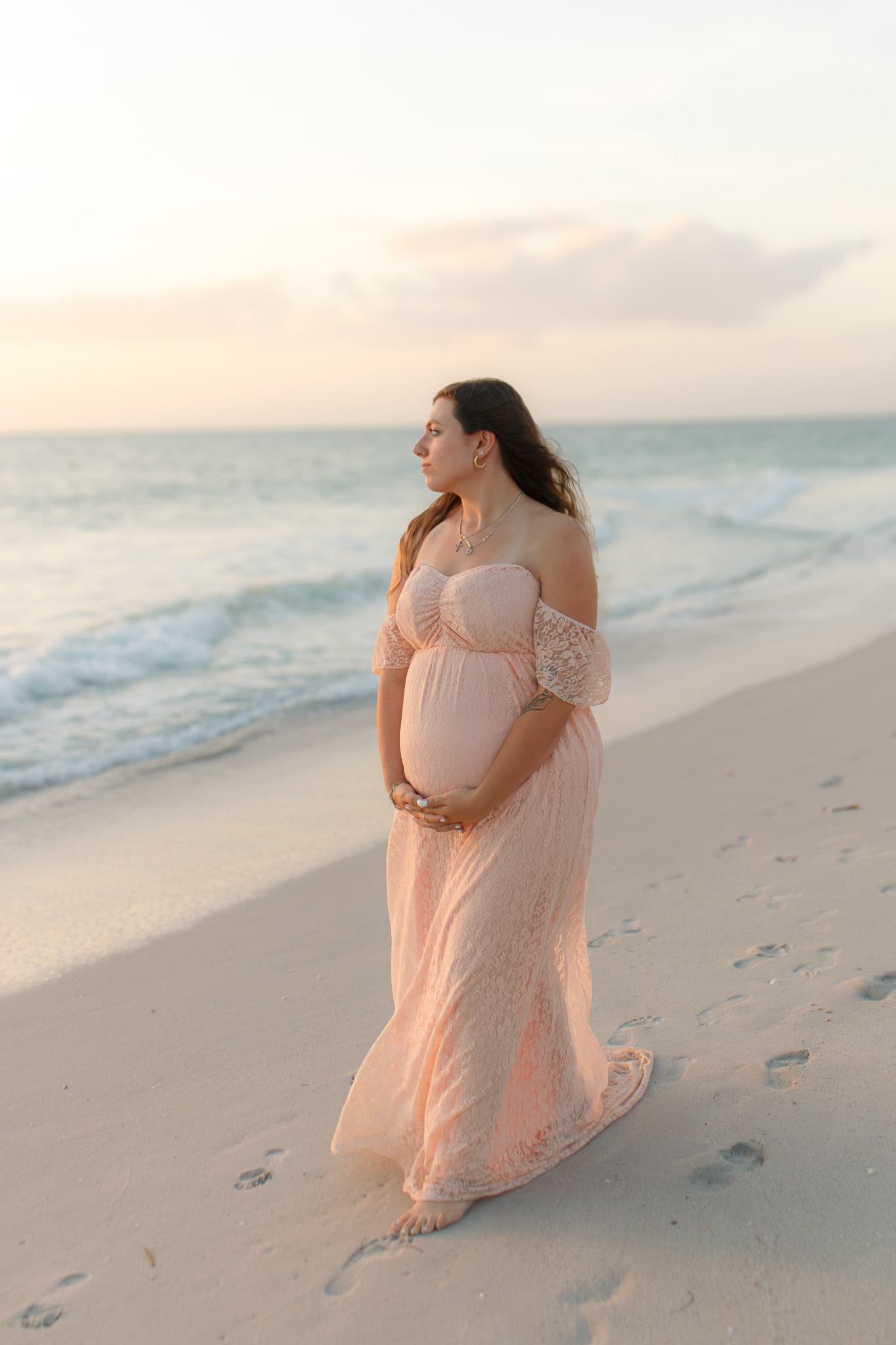 Pregnant woman in a flowing dress standing on a beach near the ocean, looking into the distance at sunset.
