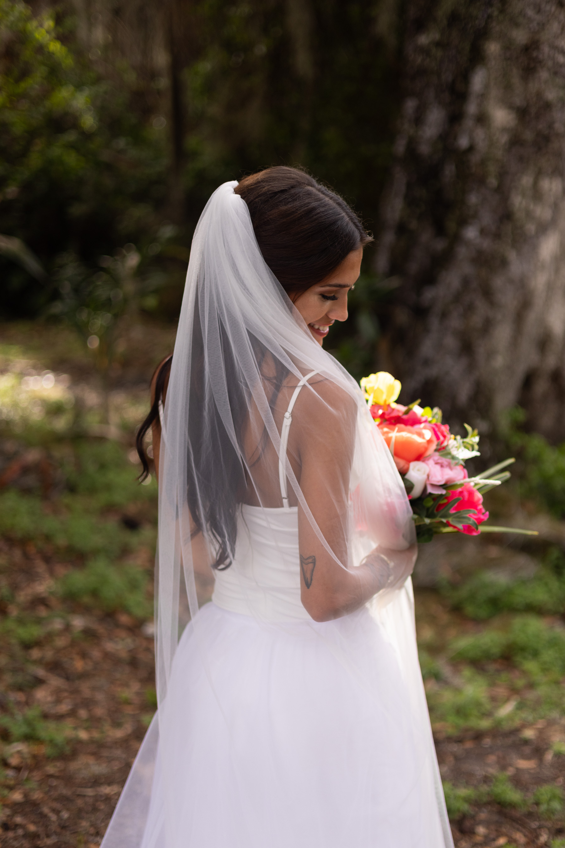 Bride in wedding dress with veil holding a bouquet outdoors, trees in background.