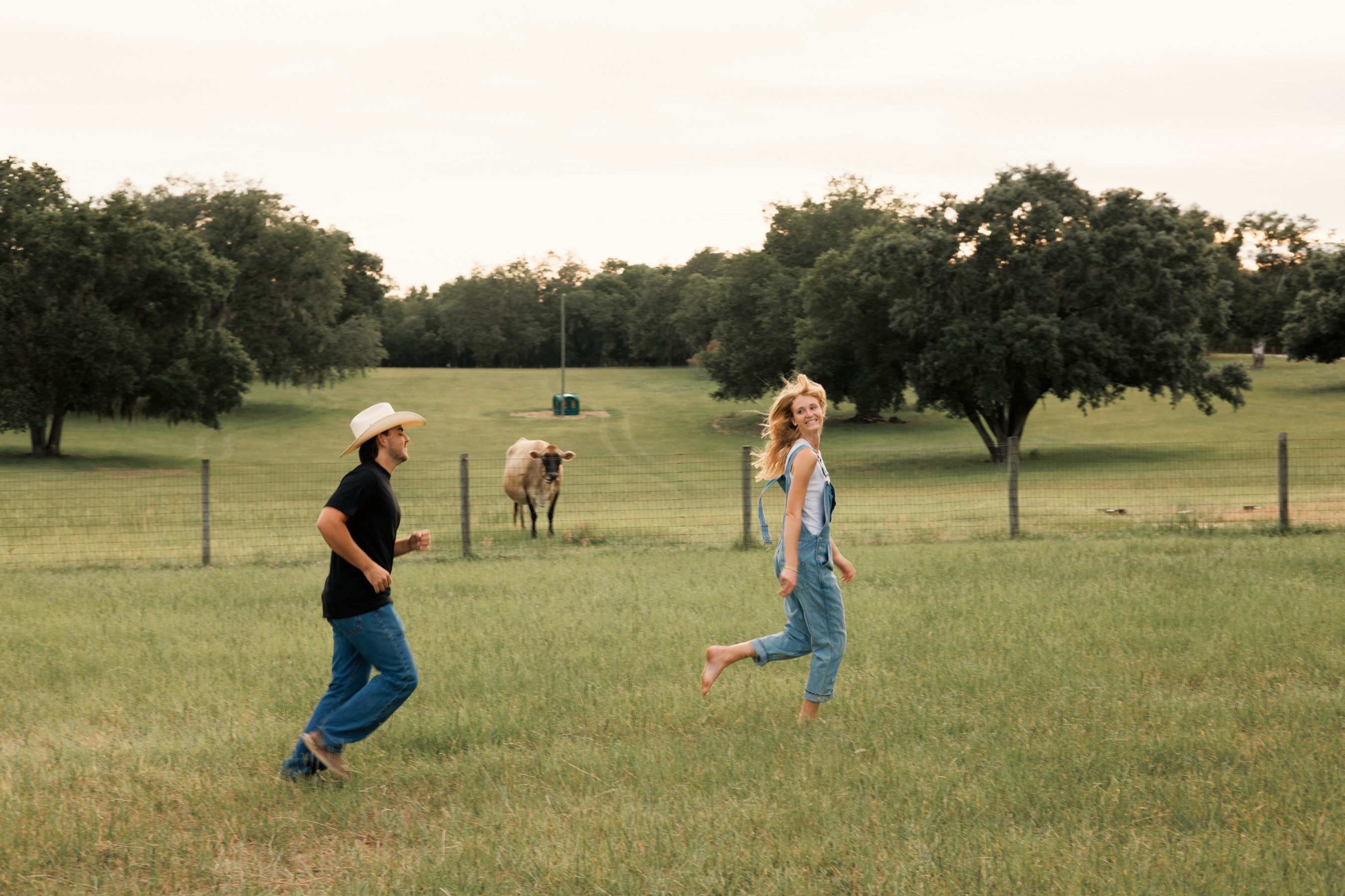 Two people playing in a grassy field with trees and a goat in the background, one person wearing a hat and the other jumping.