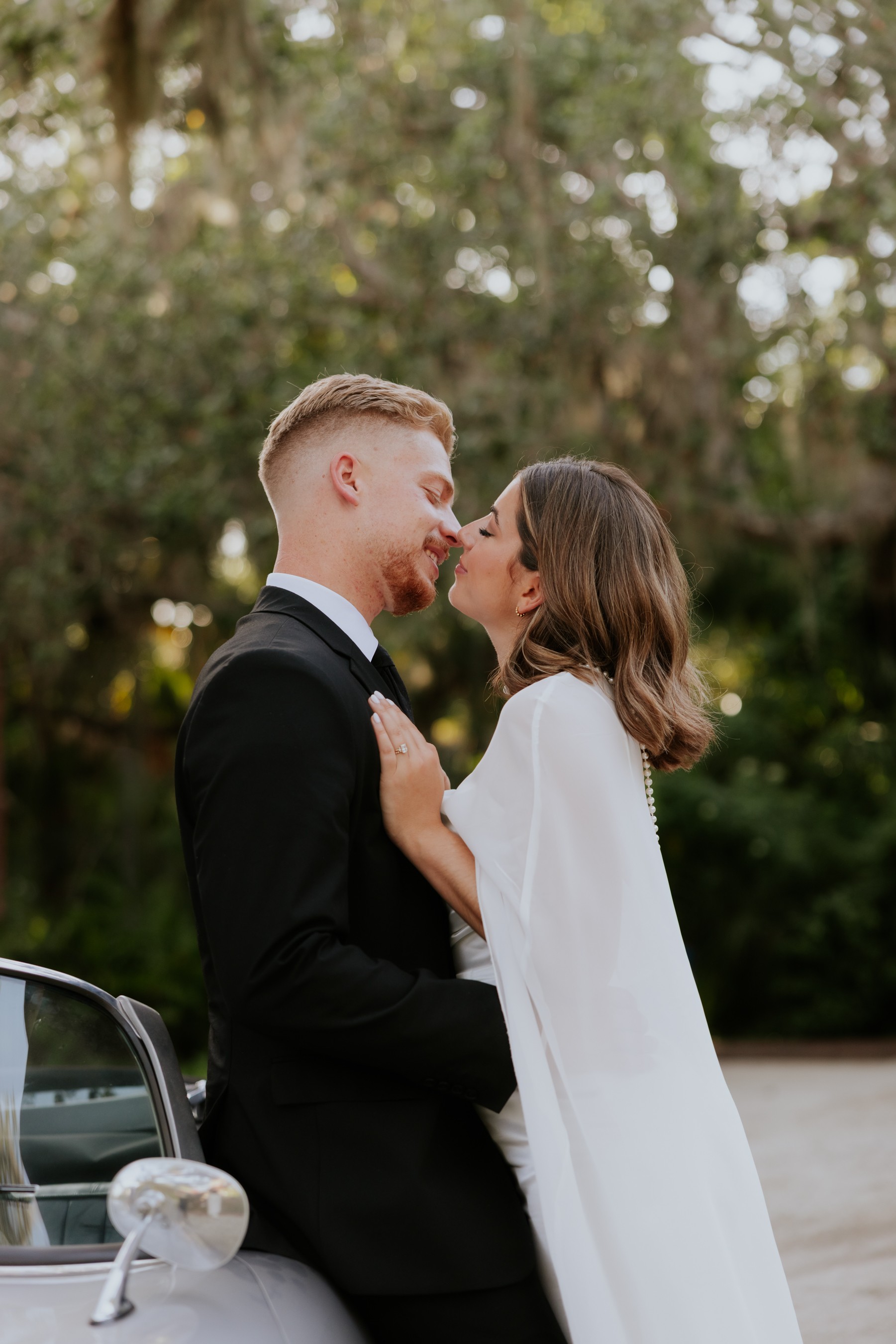 A couple in wedding attire stands close, about to kiss outdoors with trees in the background.