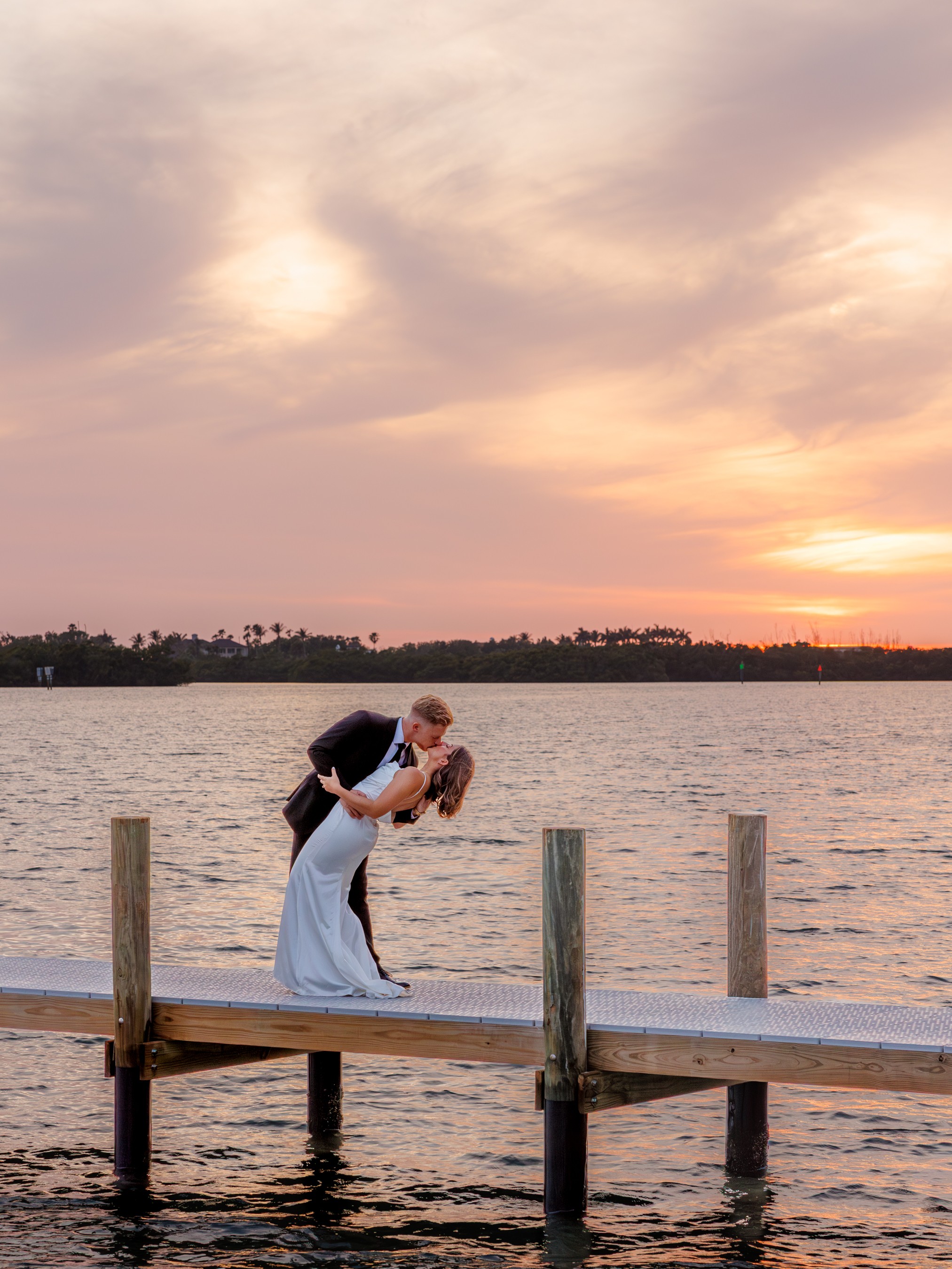 A couple stands on a wooden dock by the water during sunset, with the sky filled with soft clouds and warm colors.