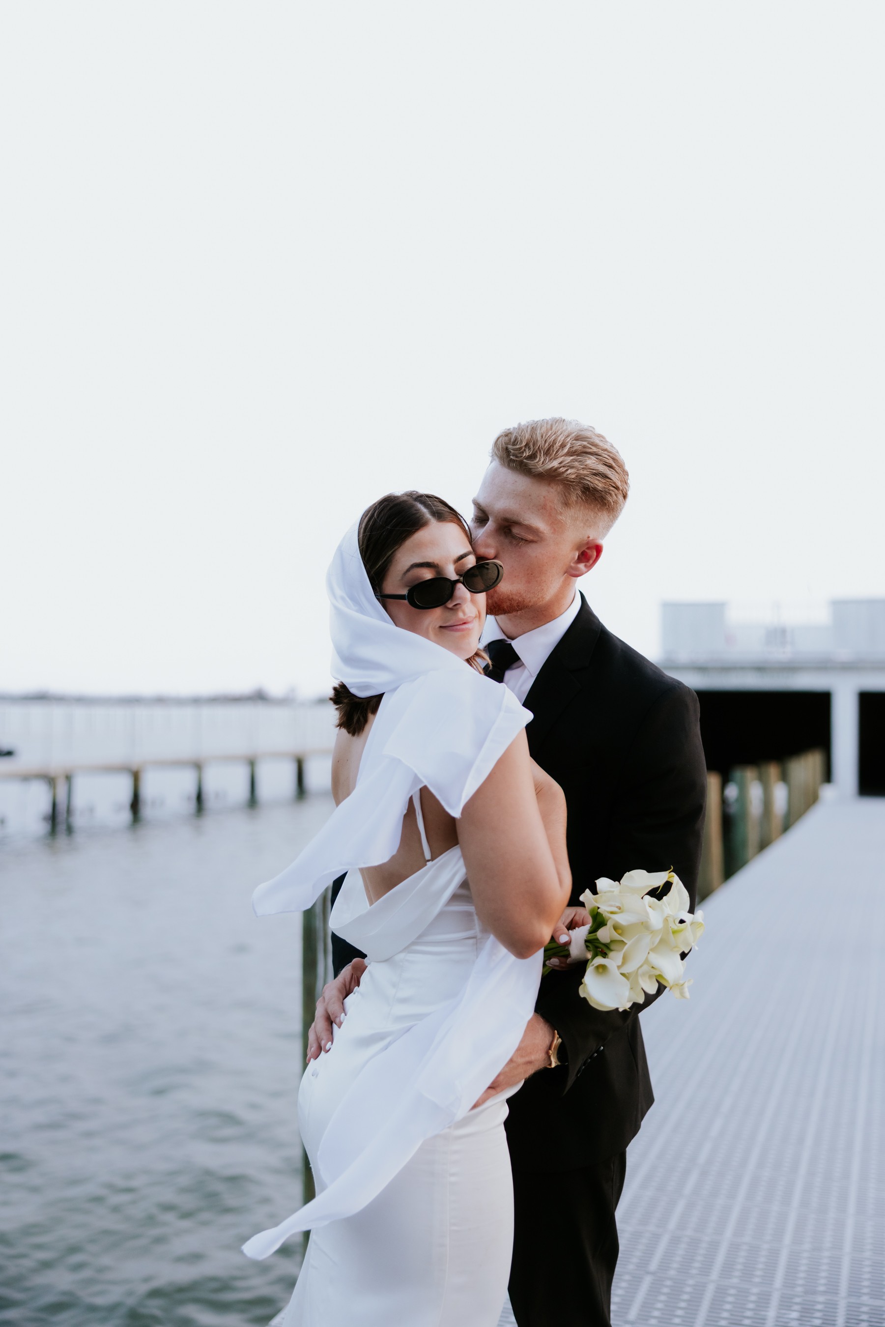 A couple dressed in wedding attire standing on a dock near water, with a cityscape in the background.