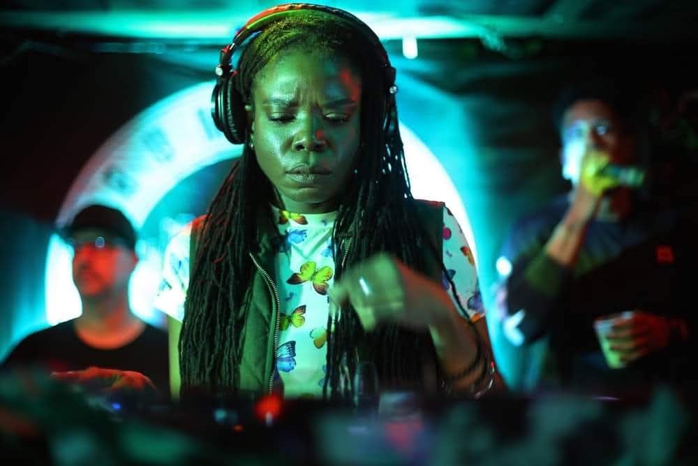 A woman with long braids wearing headphones, focused on music, with people in the background at a dimly lit event.