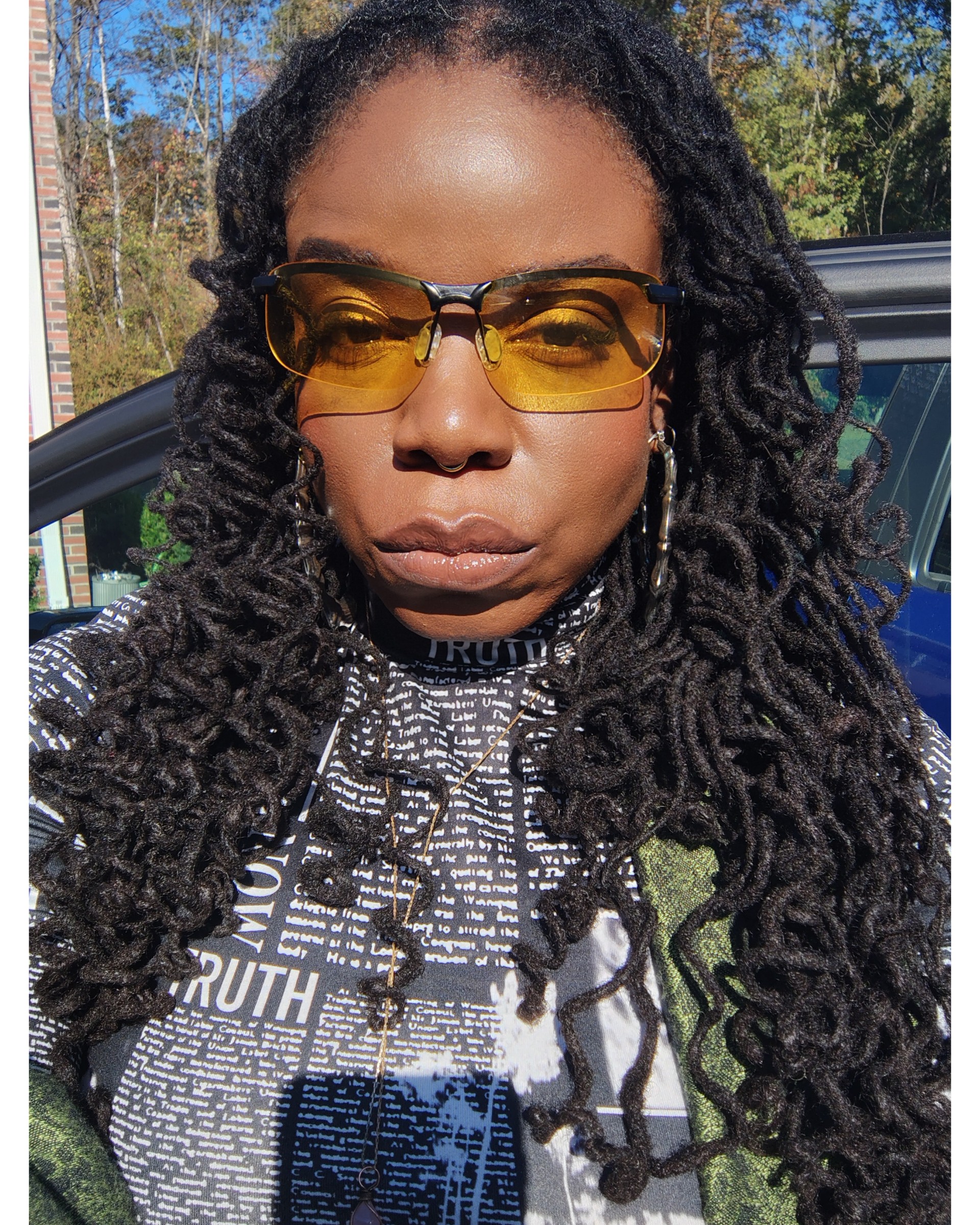 Woman with long dreadlocks wearing yellow-tinted glasses and patterned top outdoors, sunlight on face.