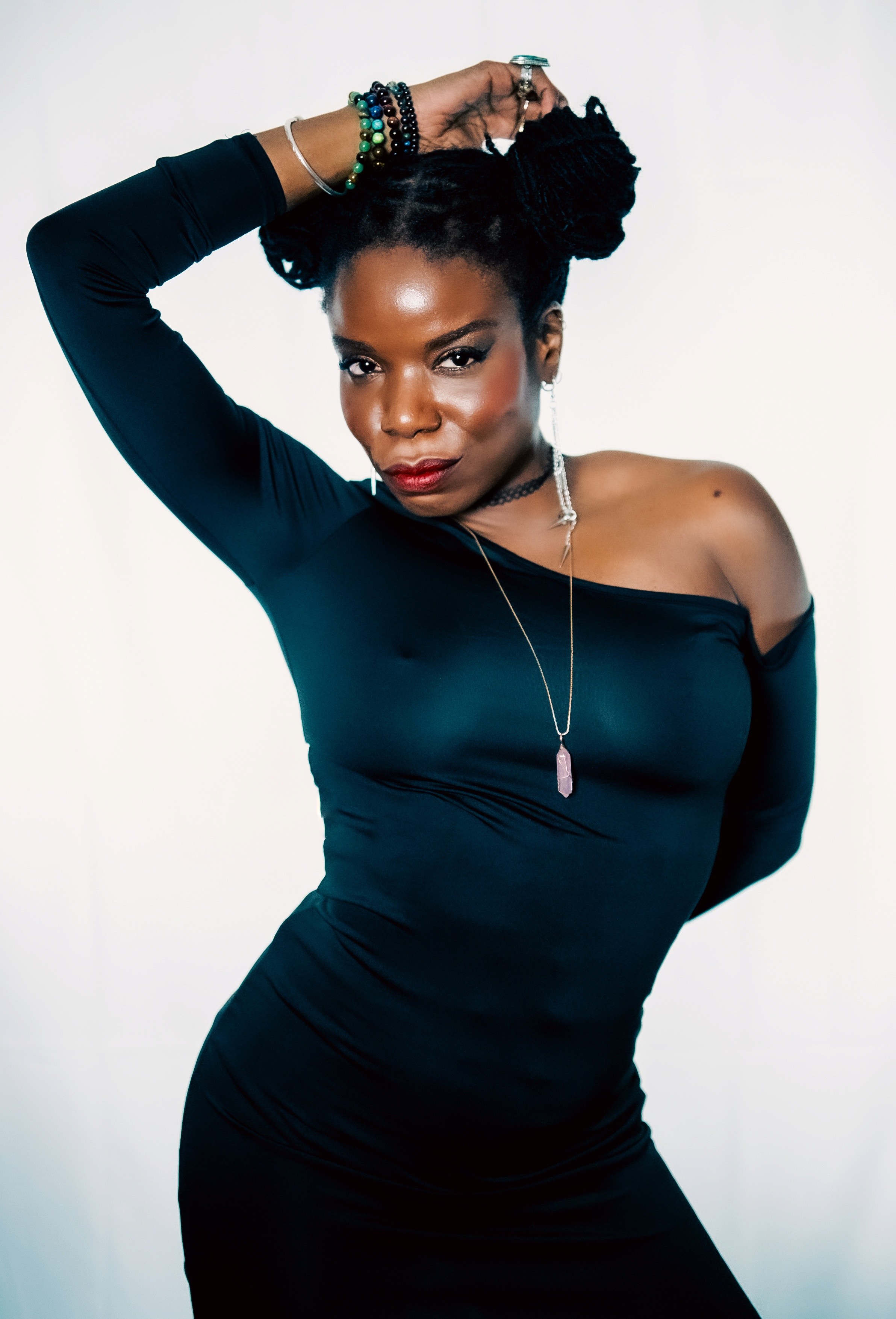 Woman with braided hair, wearing a black off-shoulder dress, jewelry, and posing with one arm raised, against a plain background.