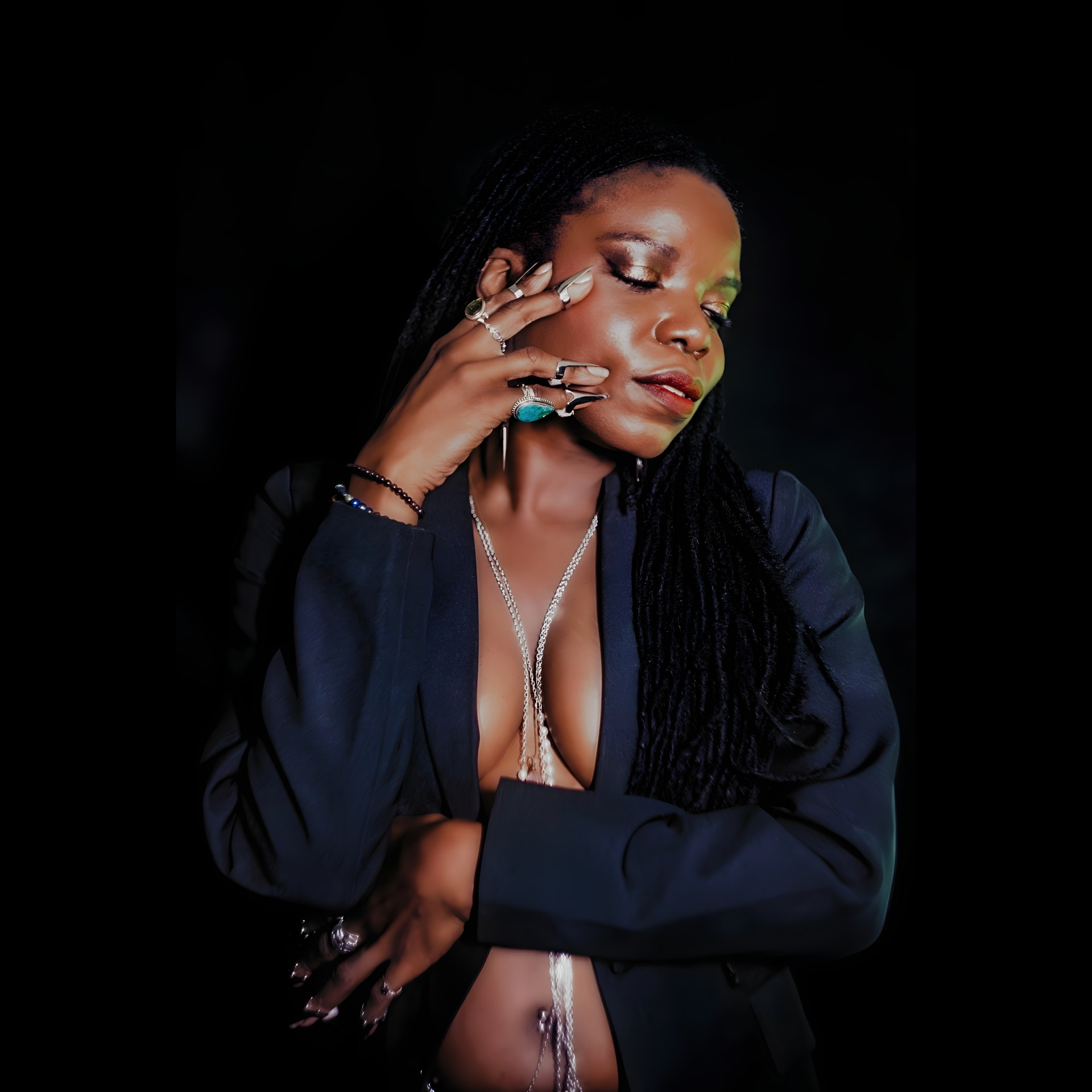Woman with long braided hair, wearing a dark blazer, jewelry, and a deep neckline, posing with her hand near her face against a black background.