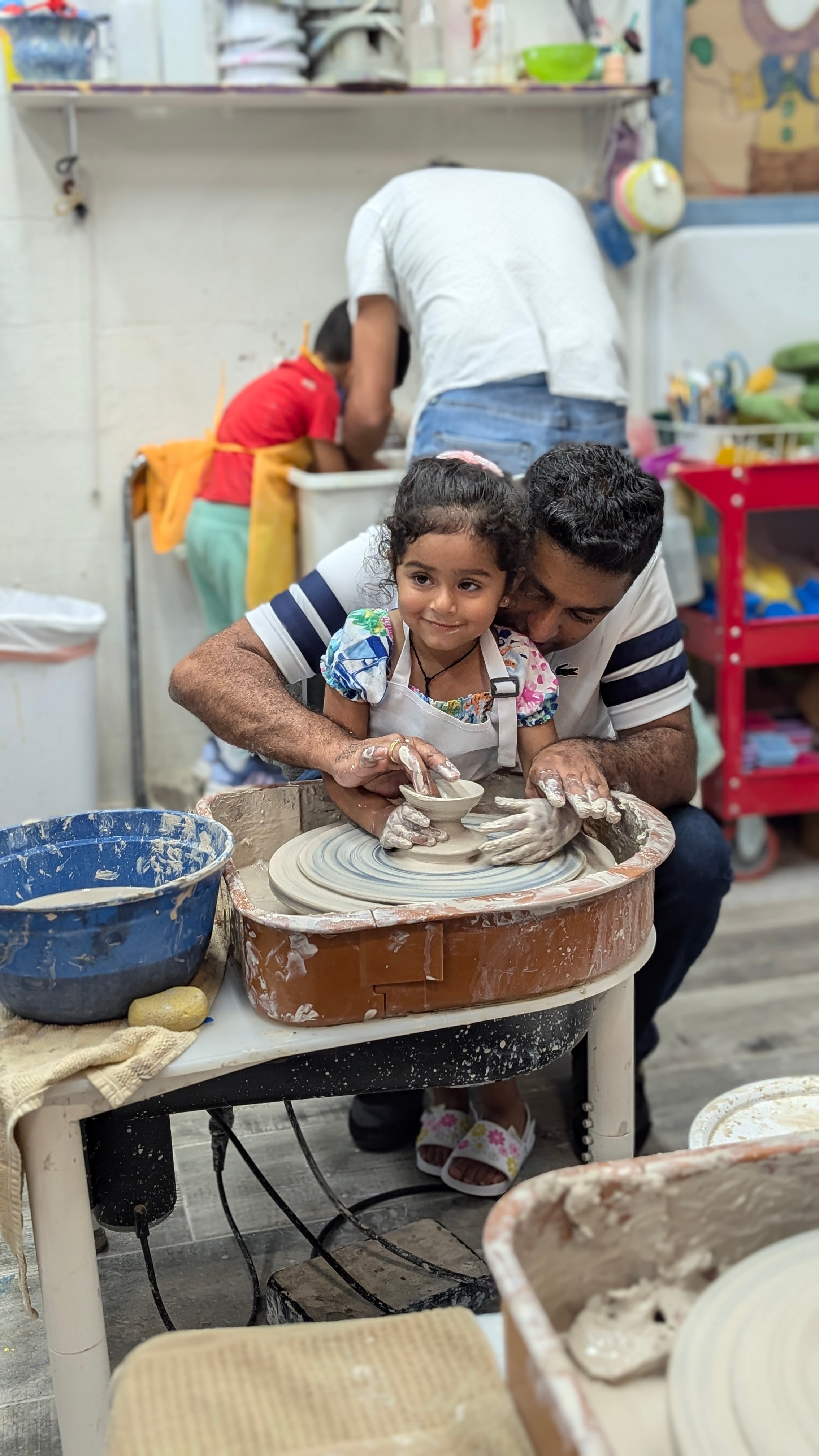 A young girl and a man are working together on a pottery wheel, shaping clay in a workshop.
