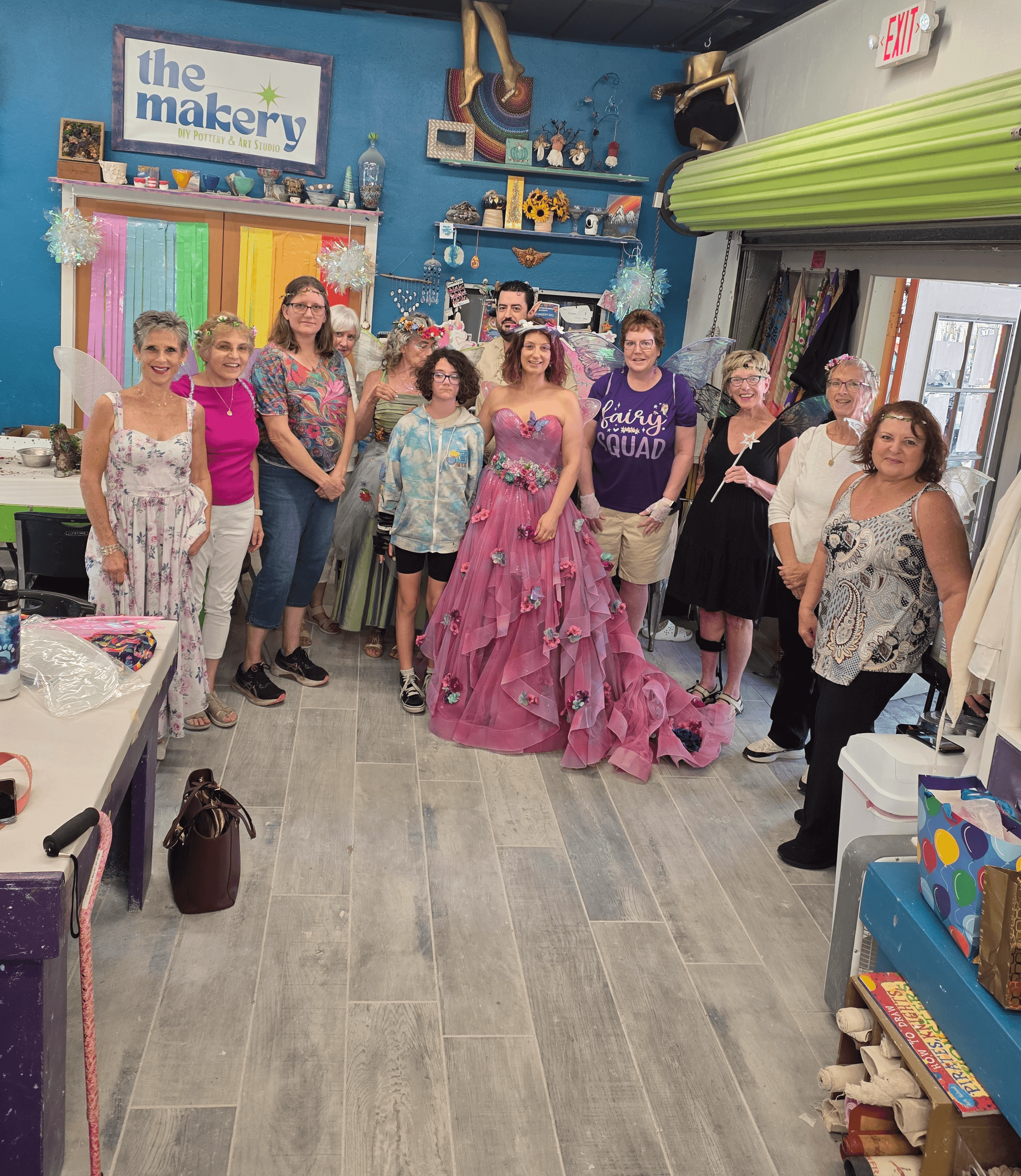 Group of women and girls standing inside a colorful craft store, some smiling and waving, with shelves and decorations in the background.