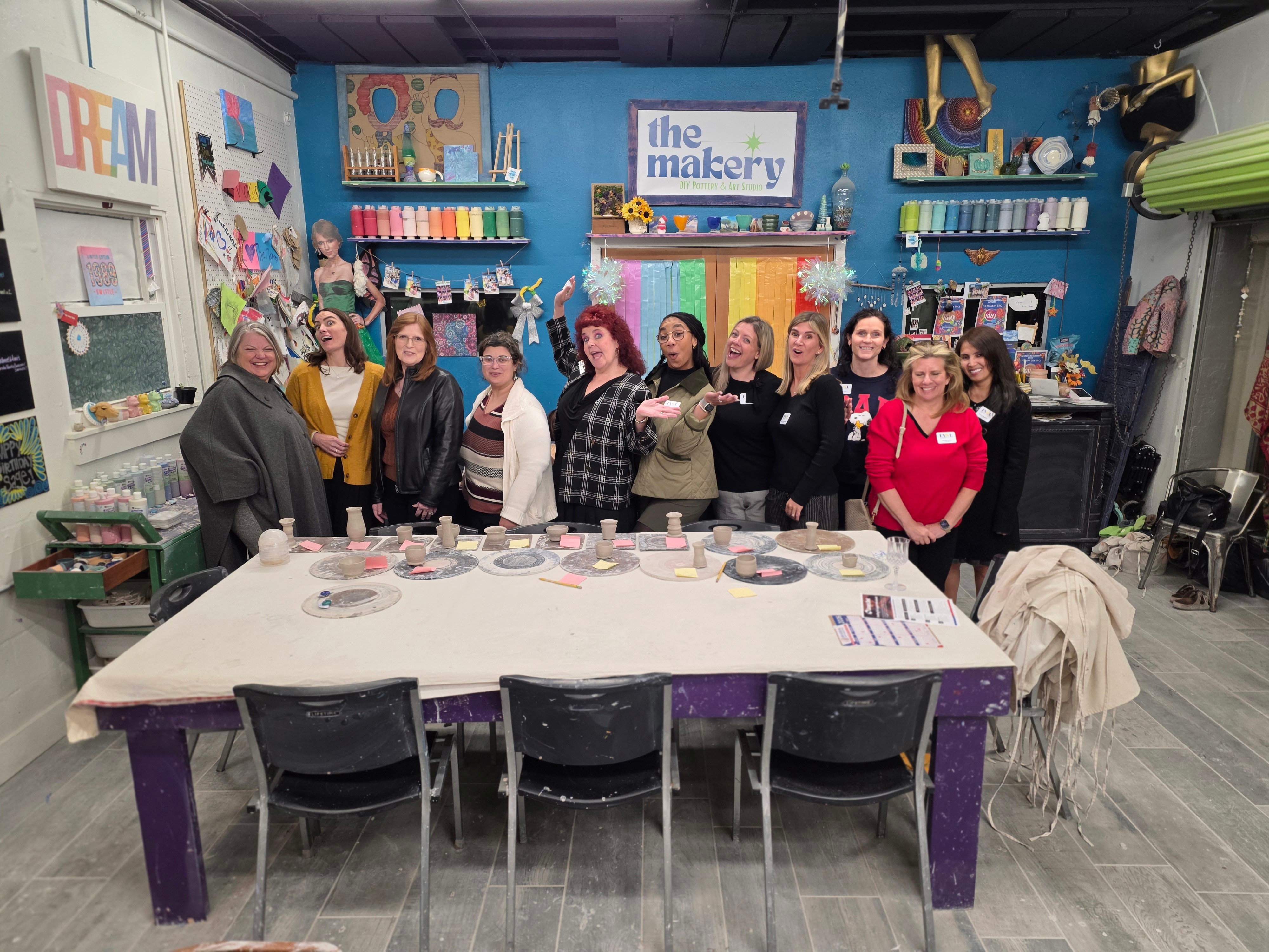 Group of people standing behind a table in a colorful craft room, with shelves and art supplies in the background.