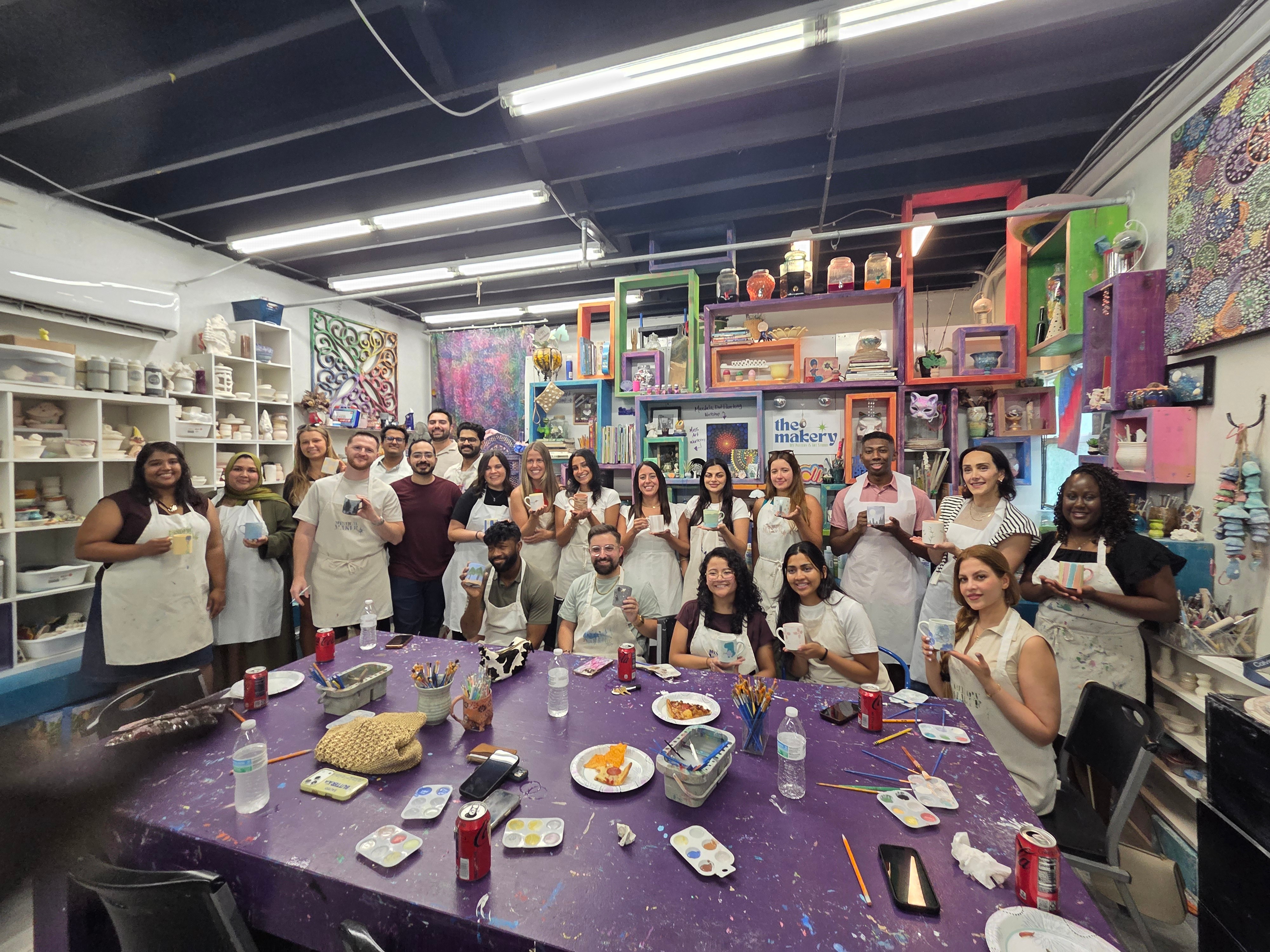 Group of people in a colorful room with shelves and tables, some holding food and drinks, smiling at the camera.