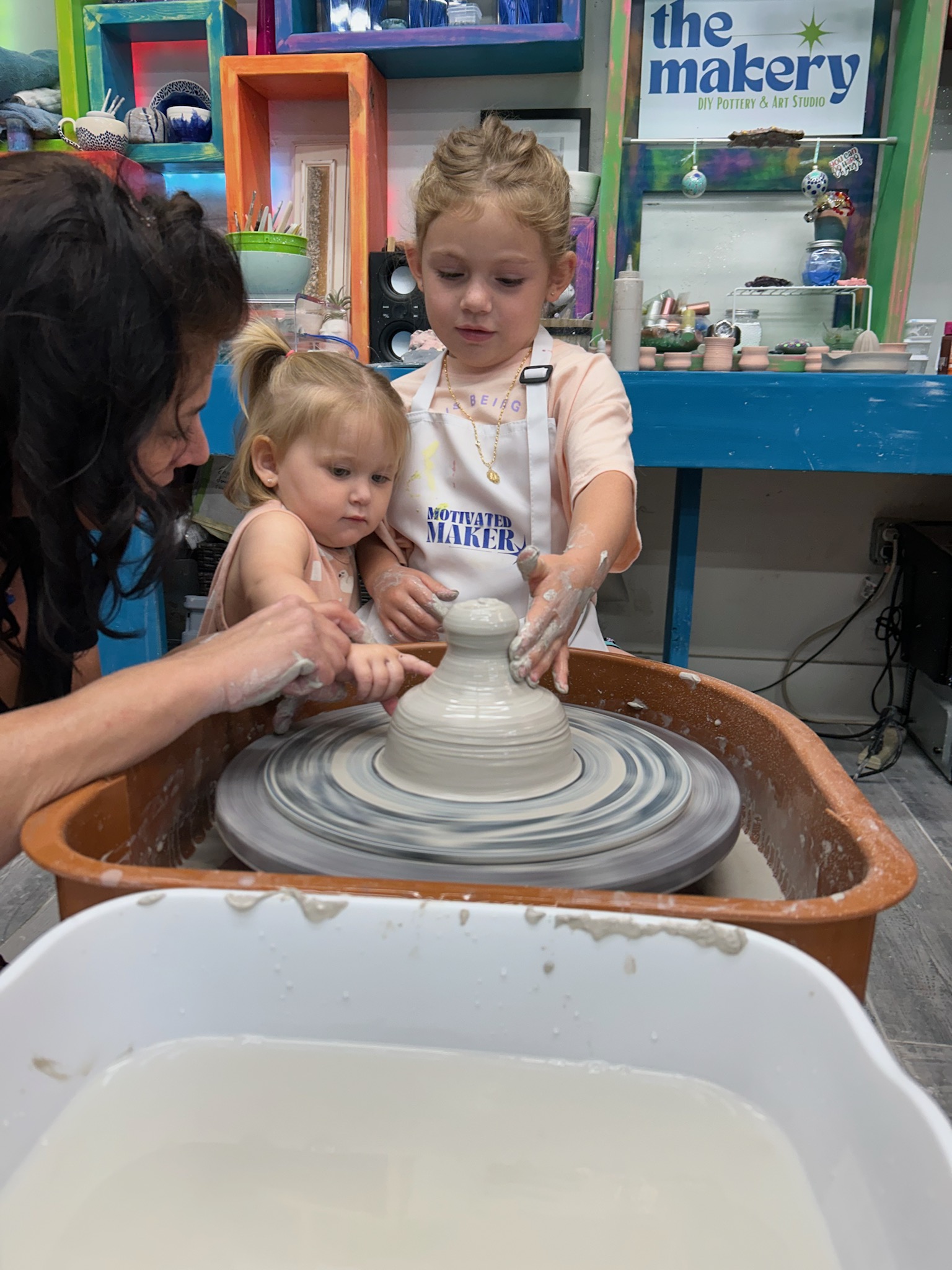 Children and an adult working on a pottery wheel in a classroom, with shelves and a sign in the background.