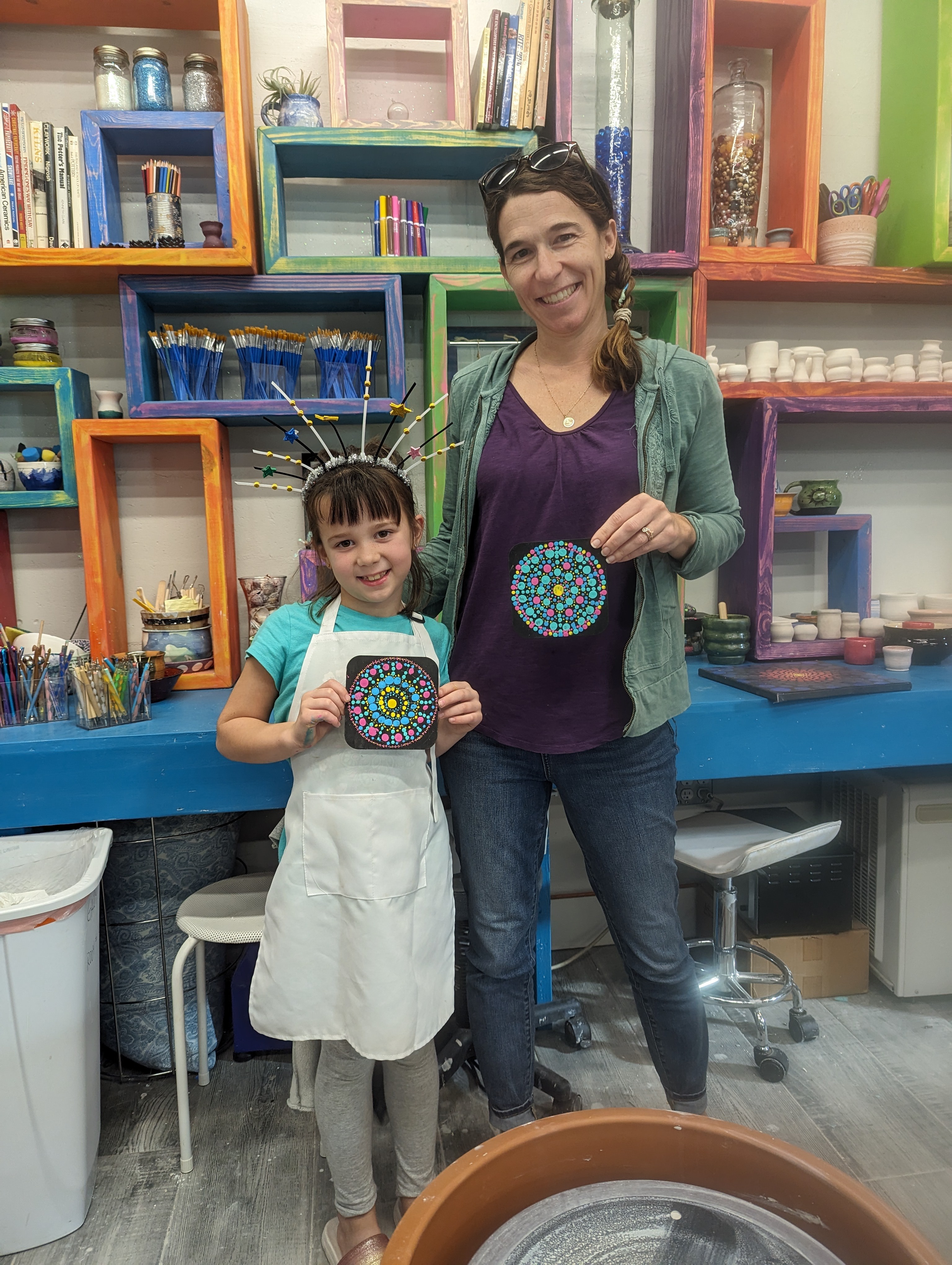 A woman and a girl stand in an art studio, holding colorful circular artwork, smiling at the camera.