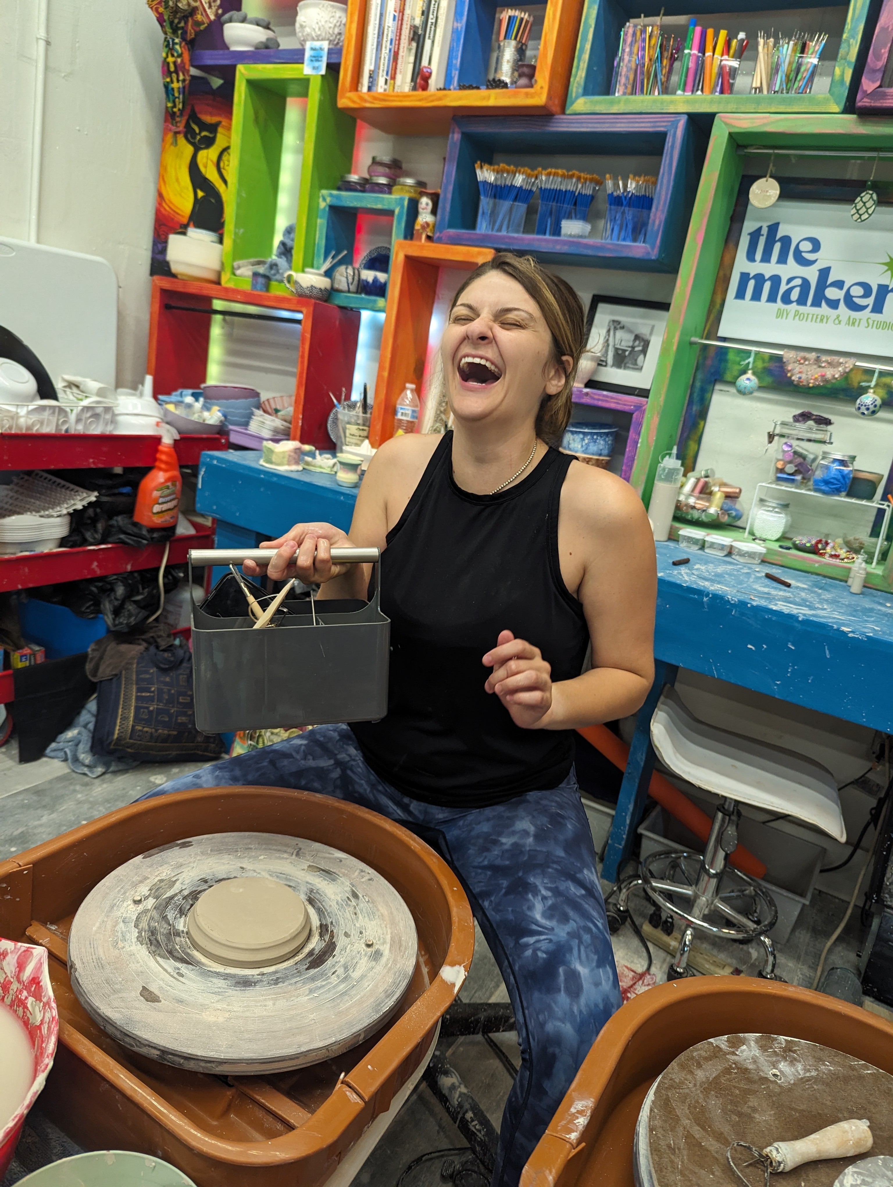 Woman laughing while working on a pottery wheel in a colorful craft room, surrounded by shelves and art supplies.