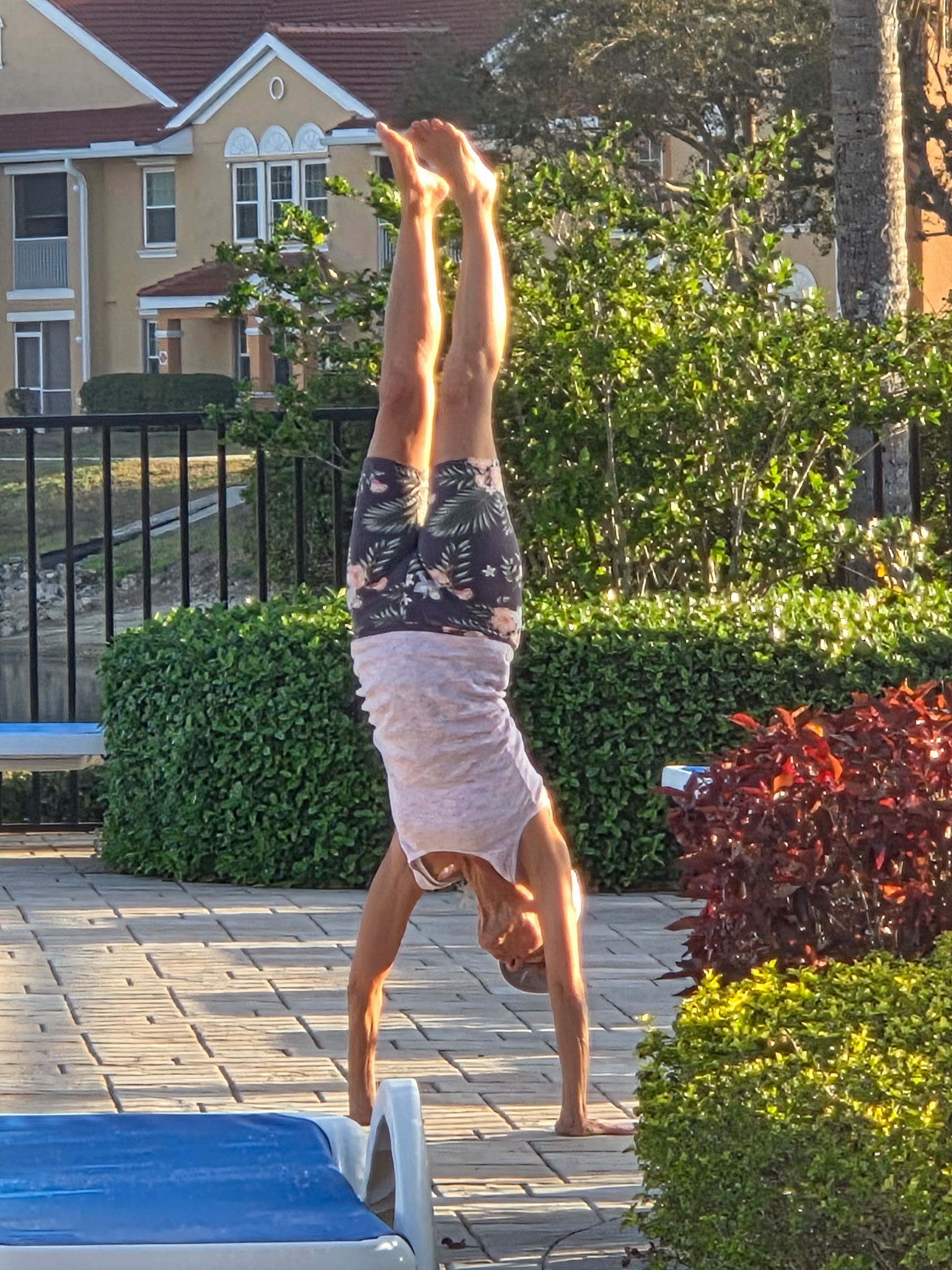 Person performing a handstand outdoors on a paved area with greenery and houses in the background.