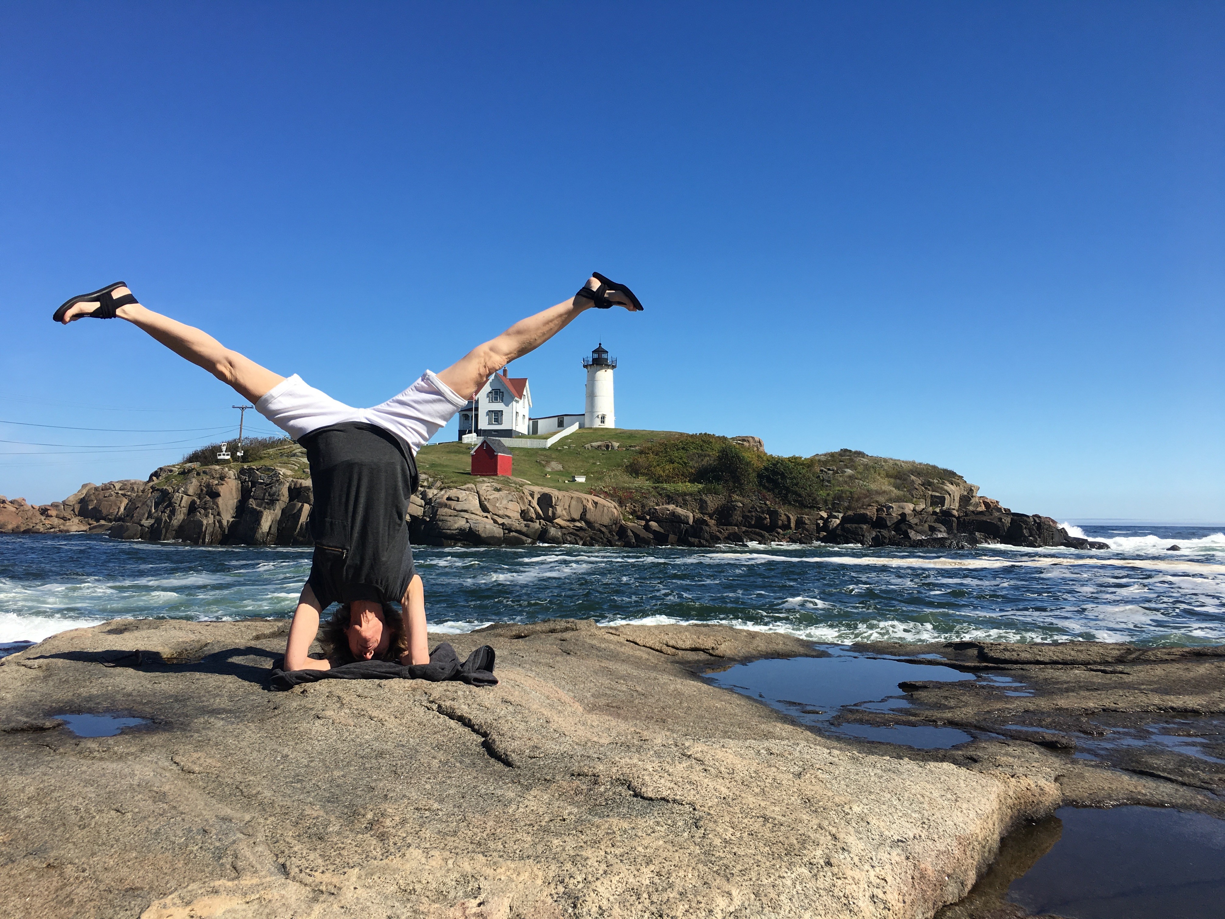 Person performing a headstand on rocks near the ocean with a lighthouse in the background under a clear blue sky.
