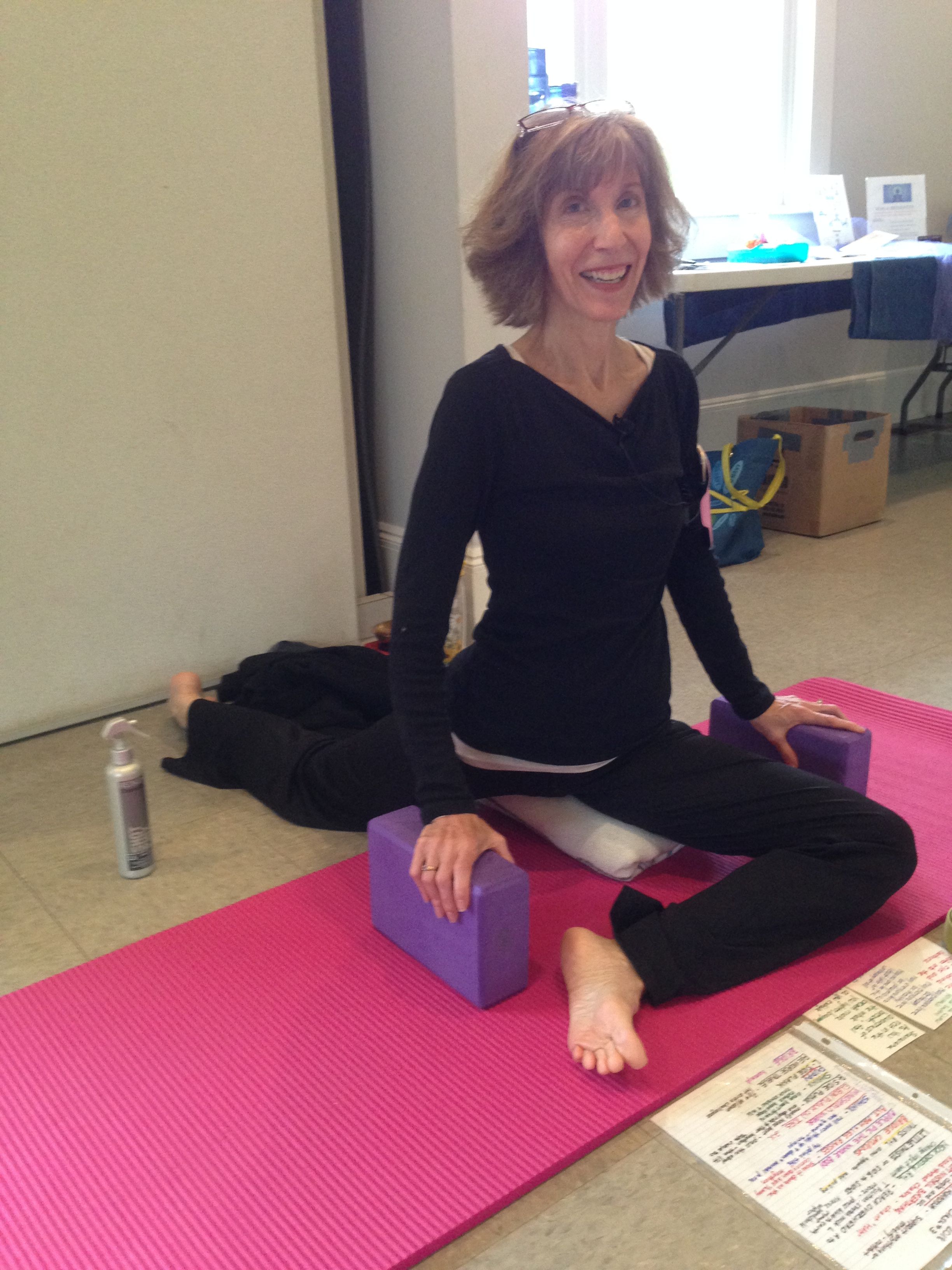 Person sitting on a pink yoga mat with a purple block, surrounded by papers and a water bottle.