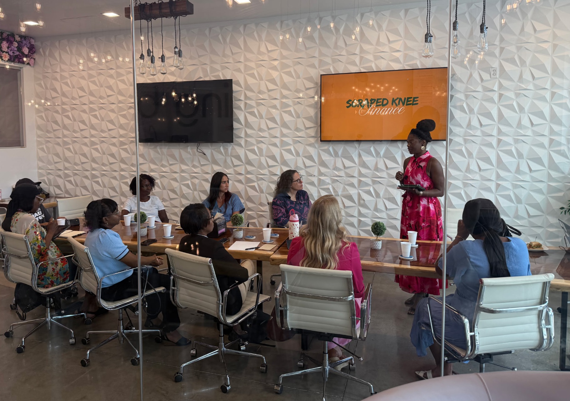 Group of people seated around a conference table listening to a woman standing and speaking, with a screen on the wall.