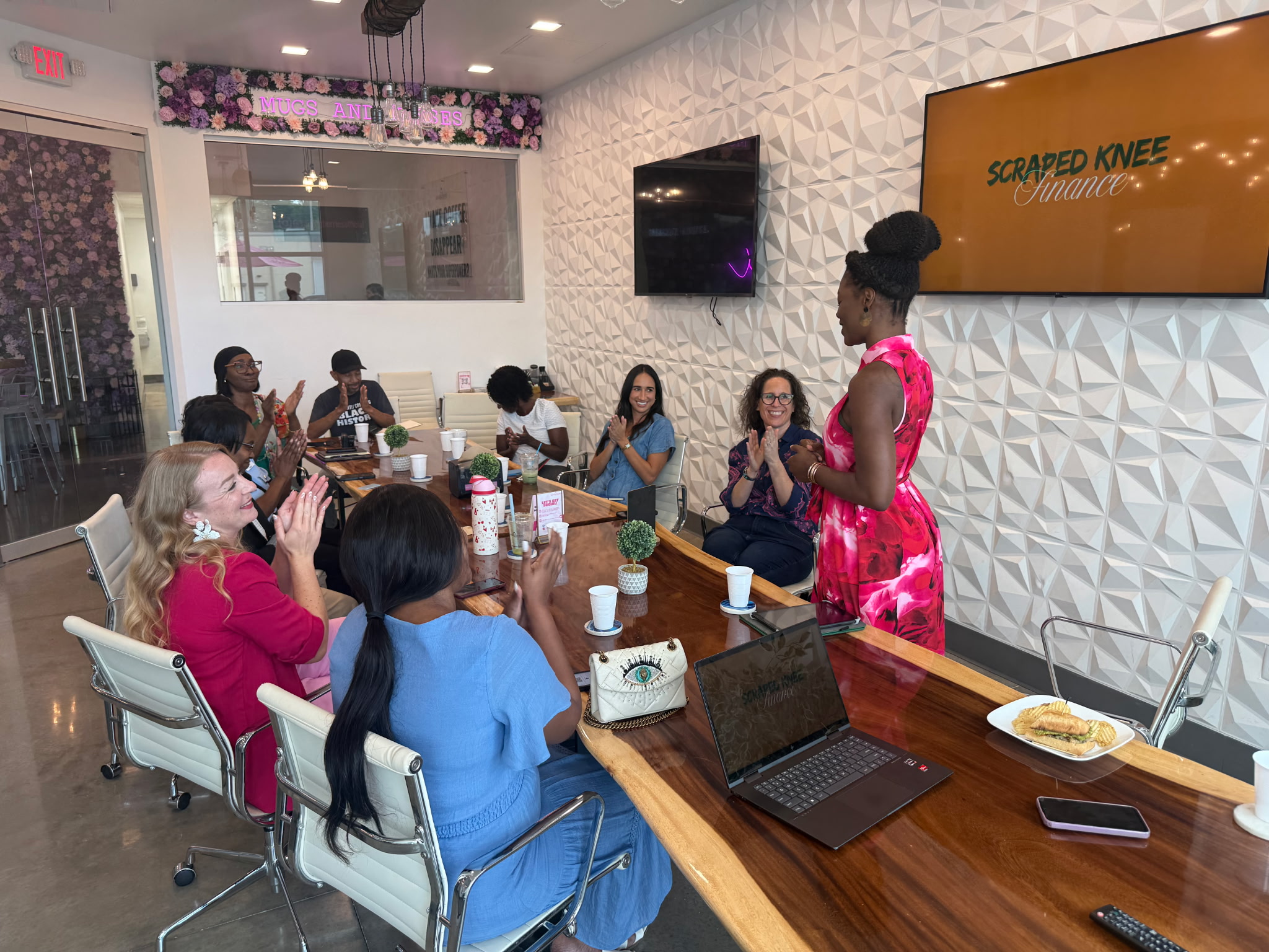 A woman in a pink dress stands in front of a group of seated women in a conference room, some taking photos.