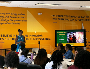 A woman presents to a seated audience in a room with yellow walls and motivational quotes, with a screen displaying a presentation.