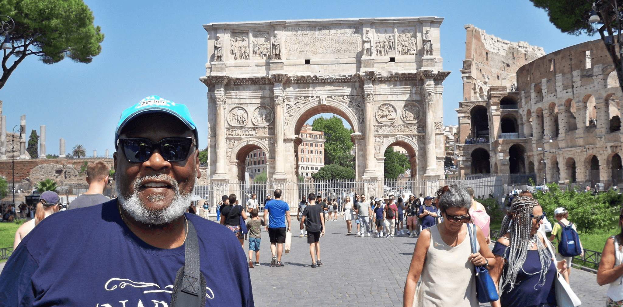 Smiling man wearing sunglasses and a cap in front of ancient Roman ruins with tourists, blue sky, and trees.