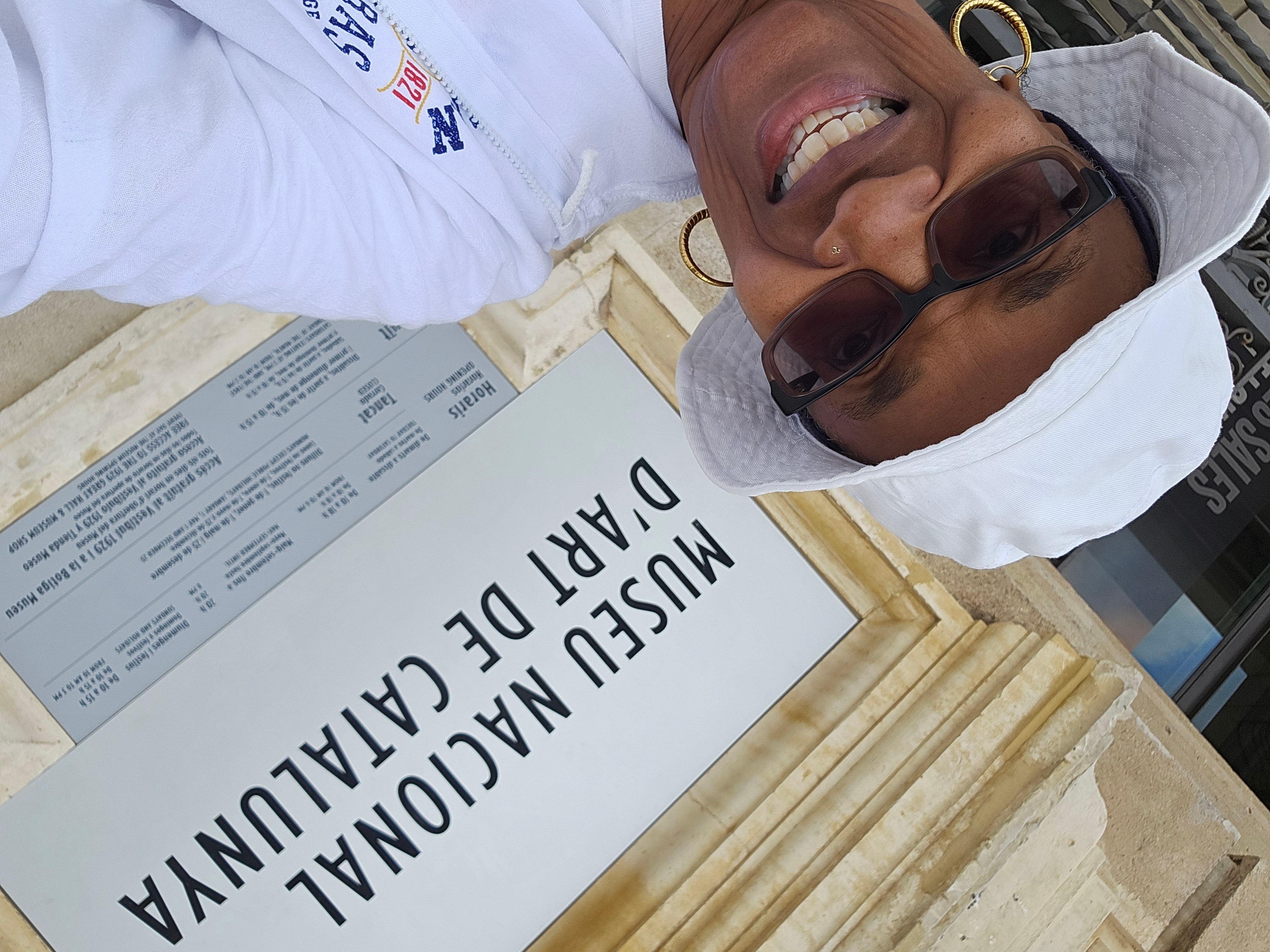 Smiling woman wearing sunglasses and a white hat, taking a selfie in front of a sign at a medical facility.