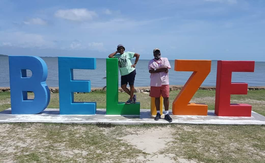 Two people stand in front of large colorful letters spelling BELIZE outdoors on a sunny day.