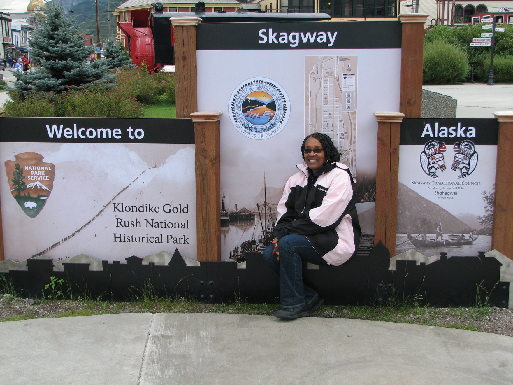 Person sitting on a bench in front of welcome signs for Klondike Gold Rush National Historical Park and Alaska, with trees and buildings in background.